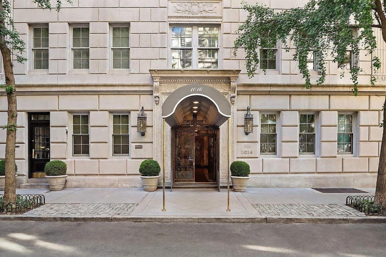 This elegant, symmetrical building facade showcases a classic limestone-style exterior with a prominent central entryway sheltered by a sophisticated fabric awning. The entrance features ornate metal doors and is flanked by symmetrical windows and decorative wall-mounted lanterns, creating a refined and prestigious street presence. Potted topiary trees add a touch of greenery to the sidewalk, while the overall composition conveys a sense of established luxury and architectural permanence.
