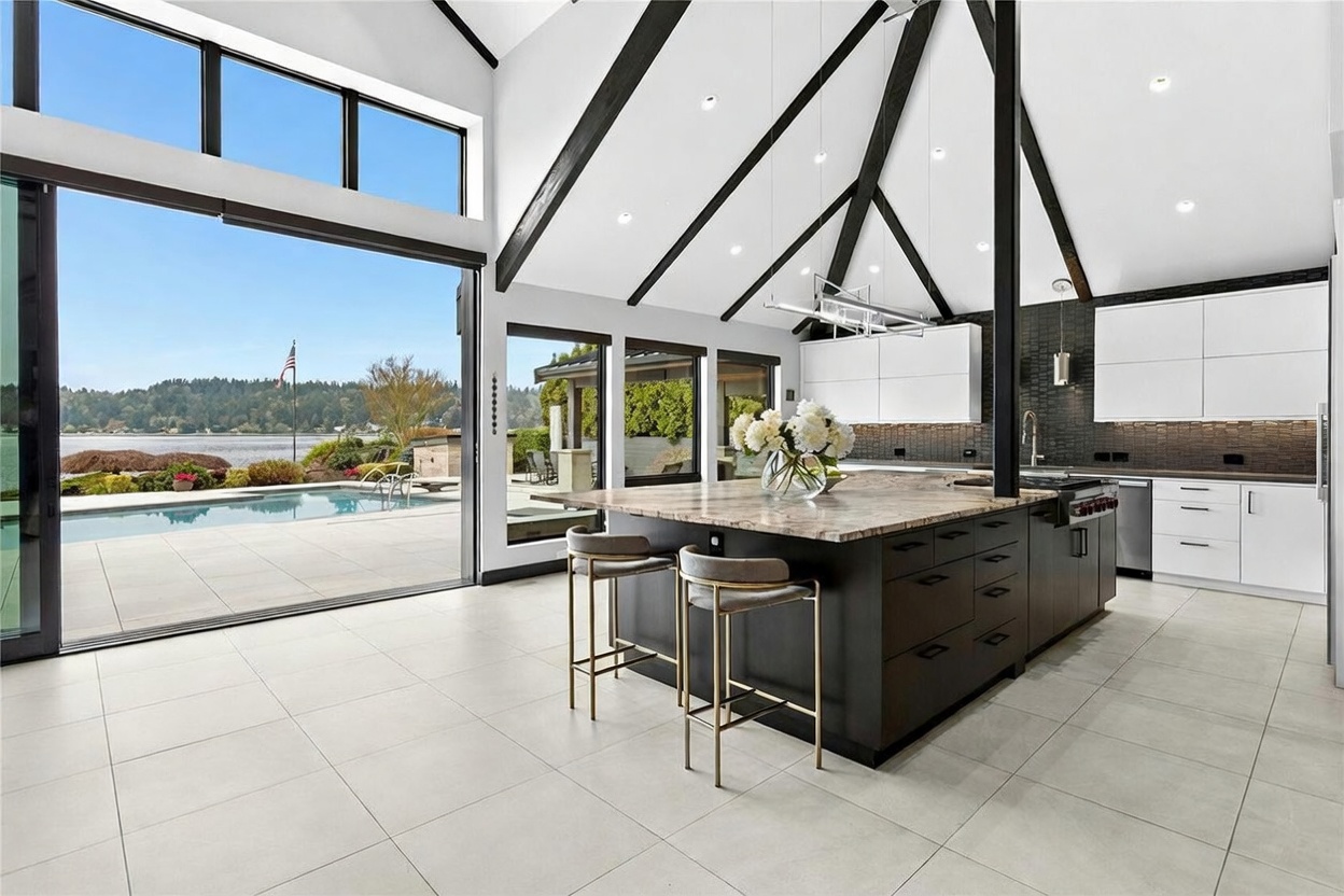 This stunning, high-end modern kitchen features a large central island with a stone countertop and gold-accented bar stools, set against a backdrop of sleek white cabinetry and dark tiled walls. The room boasts dramatic vaulted ceilings with exposed black beams and expansive floor-to-ceiling glass sliding doors that seamlessly blend the indoor space with a luxurious outdoor pool and waterfront view. The overall aesthetic is sophisticated and airy, emphasizing a seamless indoor-outdoor living experience.