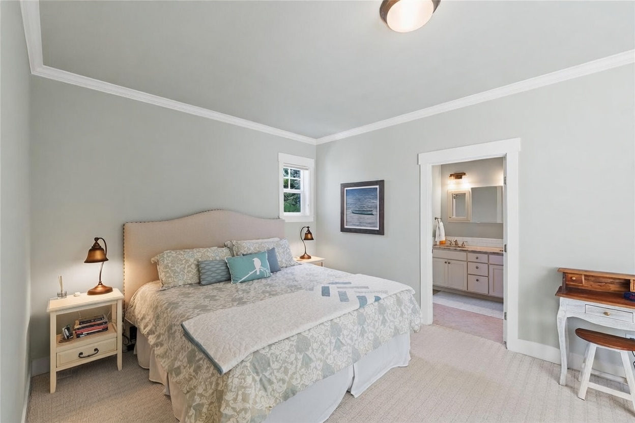 This primary bedroom features a neutral color palette with soft gray walls and a plush, light-colored carpet. A large, upholstered headboard serves as the focal point, complemented by matching nightstands with vintage-style lamps and a small desk area to the right. An open doorway provides a clear view into an adjoining bathroom, creating a cohesive and inviting suite layout.