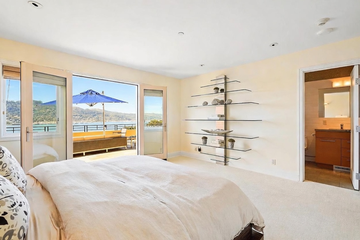 This bright primary bedroom features a neutral color palette and a large glass door that opens onto a private balcony with scenic water views. The room is furnished with a comfortable bed and a unique, tiered glass shelving unit mounted on the wall. A doorway to the right leads into an en-suite bathroom with wood-toned cabinetry, creating a cohesive and airy living space.
