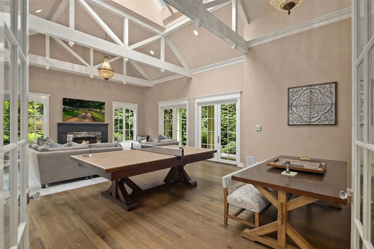 This spacious living area features a high vaulted ceiling with exposed white trusses and a large skylight, creating an airy and open atmosphere. The room is furnished with a comfortable gray sectional sofa facing a dark stone fireplace, while a ping pong table and a game table occupy the foreground. Large glass doors provide ample natural light and views of the lush greenery outside, contributing to a sophisticated and inviting recreational space.