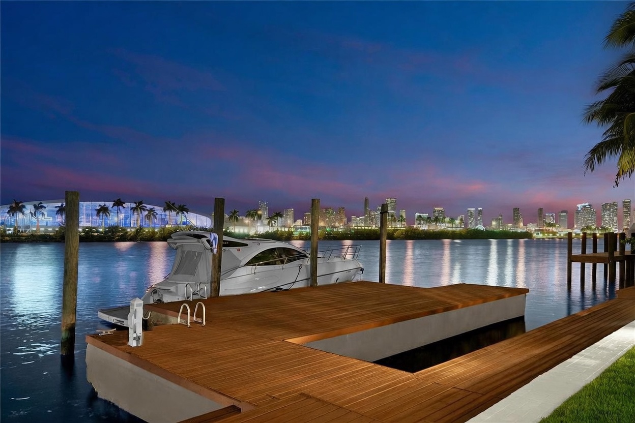 This image captures a serene waterfront deck at twilight, featuring a wooden dock extending into a calm bay. A sleek white motorboat is moored alongside the dock, while the background showcases a vibrant city skyline illuminated against a deep blue and purple sunset sky. The perspective is low and wide, emphasizing the peaceful connection between the private outdoor space and the urban horizon.