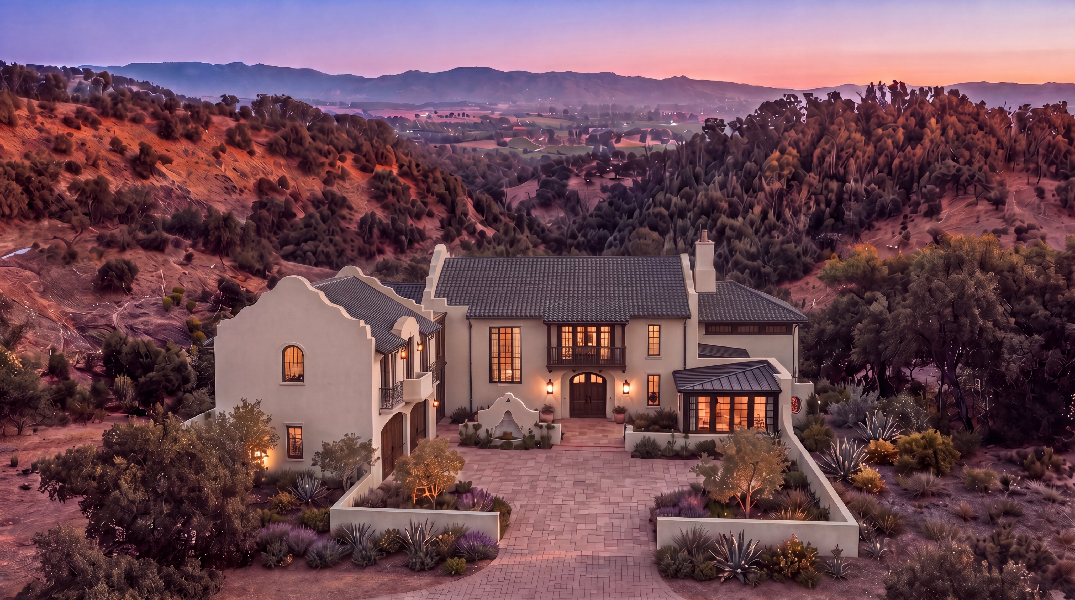 This stunning Mediterranean-style residence is nestled into a picturesque hillside, offering expansive views of a valley at sunset. The home features a symmetrical design with a stucco exterior, clay-tiled roofs, and elegant arched entryways, complemented by a stone-paved courtyard and thoughtfully landscaped terraced gardens. The warm glow of interior lighting creates an inviting atmosphere, highlighting the sophisticated architectural character of the property.