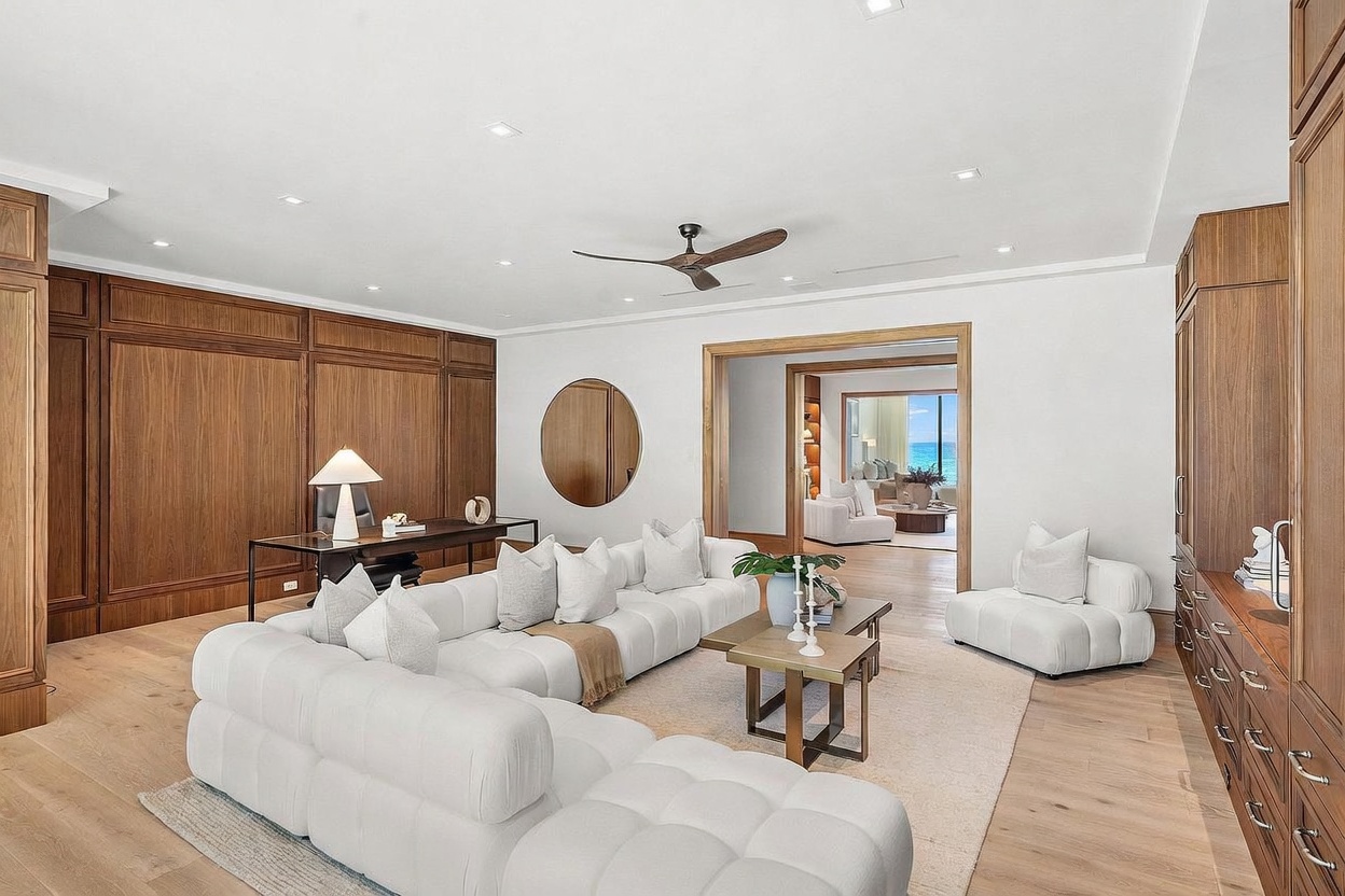 This sophisticated living room features a large, modular white sectional sofa arranged on a neutral area rug, complemented by a matching lounge chair and a pair of tiered coffee tables. The space is anchored by a wall of rich, wood-paneled wainscoting and a sleek desk area, while a circular mirror adds a modern touch. The room flows seamlessly into an adjacent space with a view of the ocean, creating an open and airy atmosphere with a refined, contemporary aesthetic.
