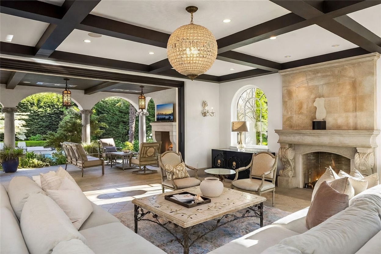 This elegant living room features a coffered ceiling with dark beams and a large crystal chandelier, seamlessly transitioning to an expansive covered patio through a wide opening. The space is anchored by a grand stone fireplace with an ornate mantel and a central seating area with a patterned area rug and a light-toned coffee table. The open-concept design creates a sophisticated indoor-outdoor flow, highlighting the lush greenery visible in the background.
