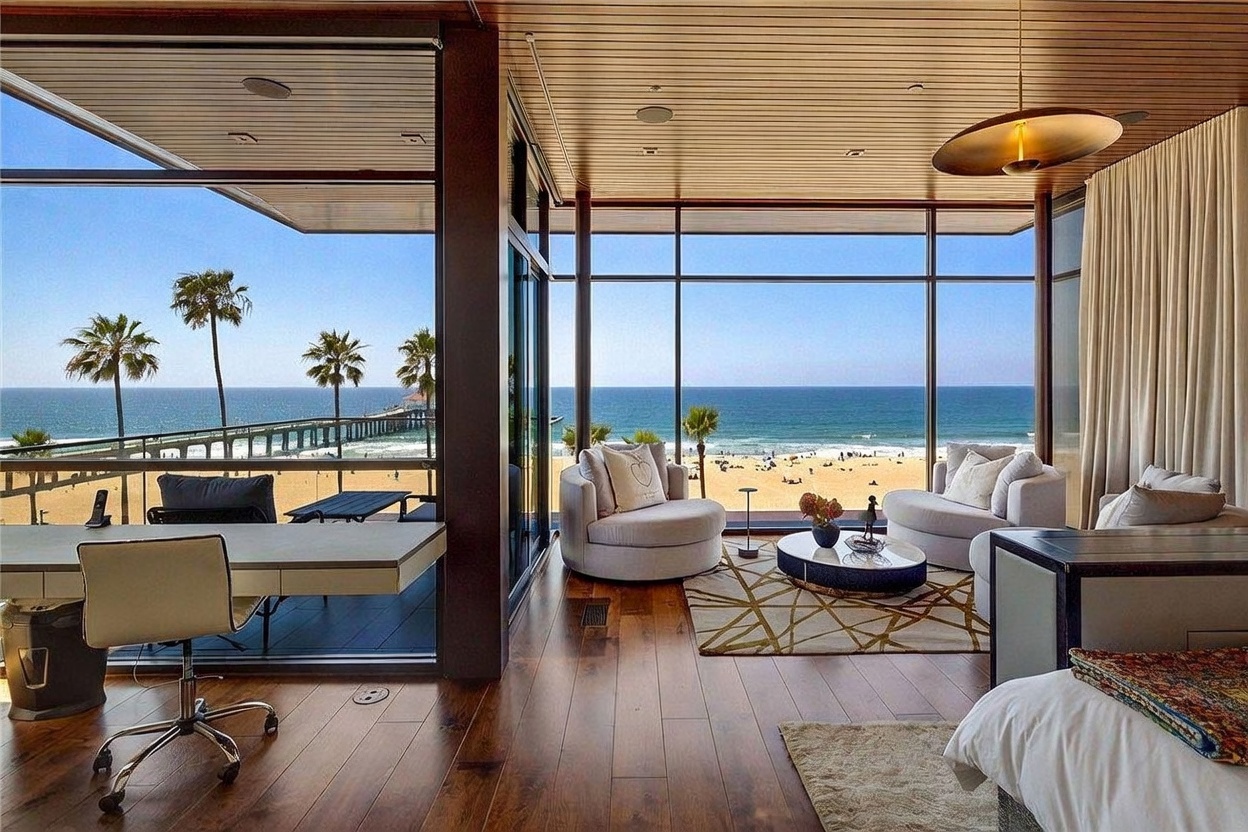 This expansive primary bedroom features floor-to-ceiling windows that offer panoramic, unobstructed views of the ocean and a nearby pier. The room is furnished with a comfortable seating area, a desk workspace, and a bed, all set against rich wood flooring and a slatted wood ceiling. The open-concept design seamlessly blends indoor luxury with the coastal landscape, creating a serene and sophisticated atmosphere.