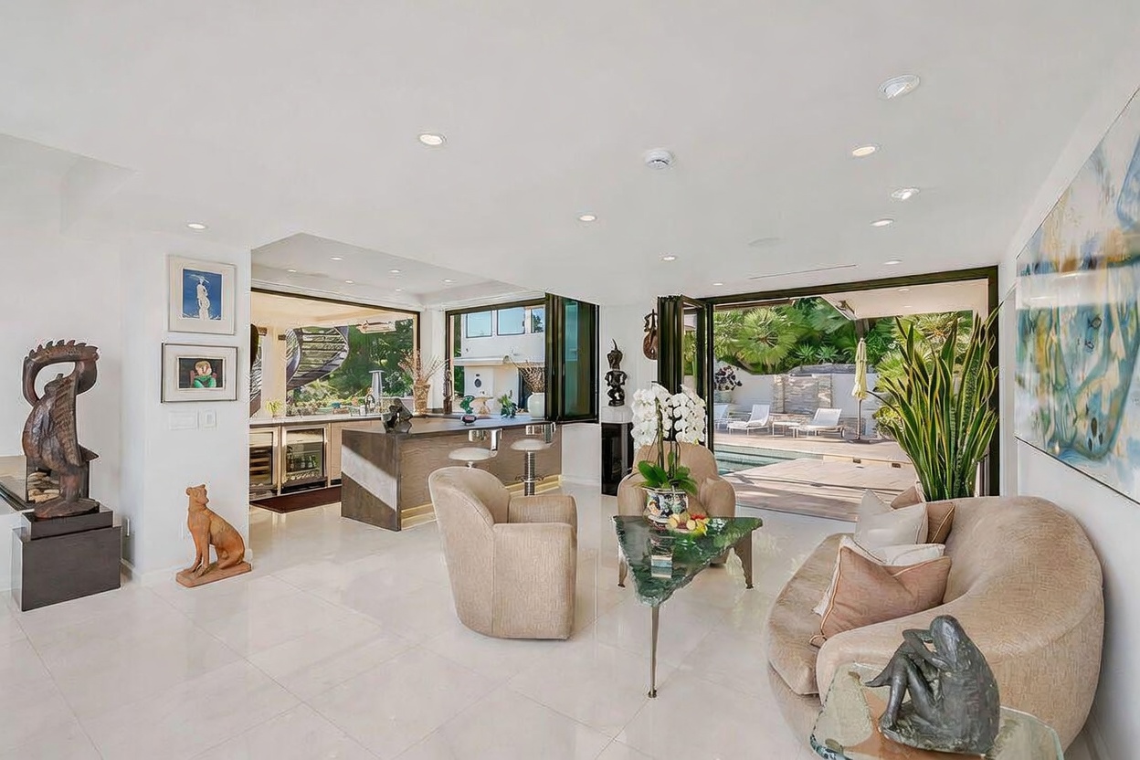 This bright, open-concept living area features a seamless transition to an outdoor pool deck through large, folding glass doors. The space is furnished with a curved sofa, a pair of armchairs, and a unique triangular glass-topped coffee table, all set against a backdrop of white walls and polished light-colored flooring. A bar area with stools and a wine refrigerator is visible in the background, creating a sophisticated space for entertaining.