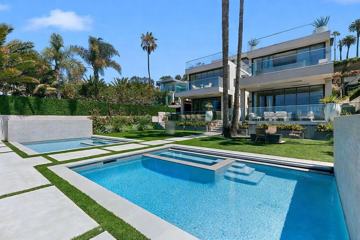 This luxurious backyard features a modern swimming pool with an integrated spa, set against a backdrop of a multi-level contemporary residence. The outdoor space includes a manicured lawn, stone pavers, and a sleek water feature, all framed by lush palm trees and a tall privacy hedge. The home showcases expansive glass walls and multiple balconies, creating a seamless transition between indoor and outdoor living areas.