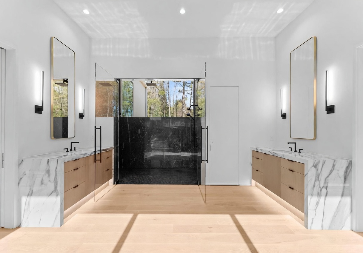 This primary bathroom features a symmetrical layout with dual vanities flanking a central glass-enclosed shower. The space is defined by light wood cabinetry, stone countertops with matching backsplashes, and tall, thin mirrors accented by modern wall-mounted light fixtures. The shower area is enclosed by glass panels and features dark, patterned tile walls, creating a striking contrast against the bright, minimalist aesthetic of the room.