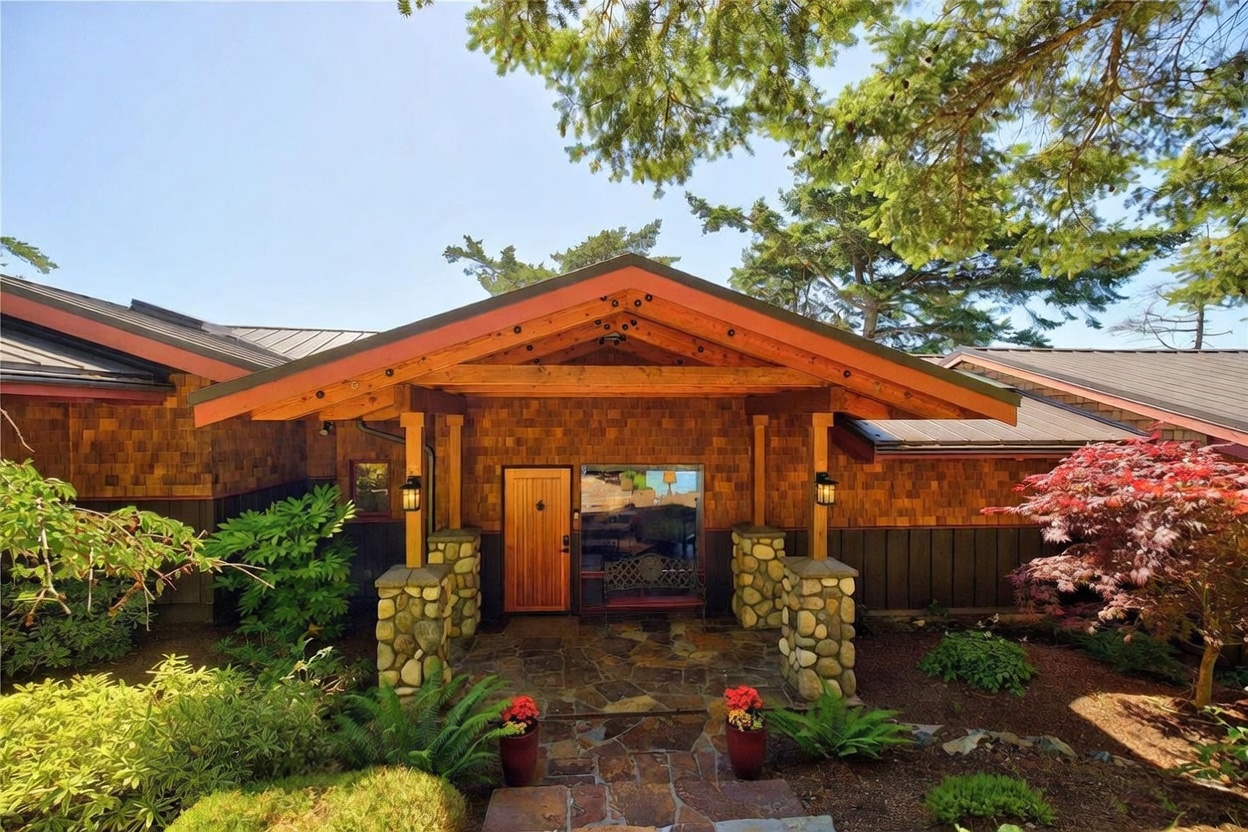 This inviting entryway features a prominent covered porch with heavy timber framing and stone-clad support pillars. The exterior is finished in warm-toned wood shingles, complemented by a natural stone walkway and lush landscaping including ferns and a vibrant Japanese maple. A wooden front door sits centered under the gable roof, creating a welcoming and rustic aesthetic.