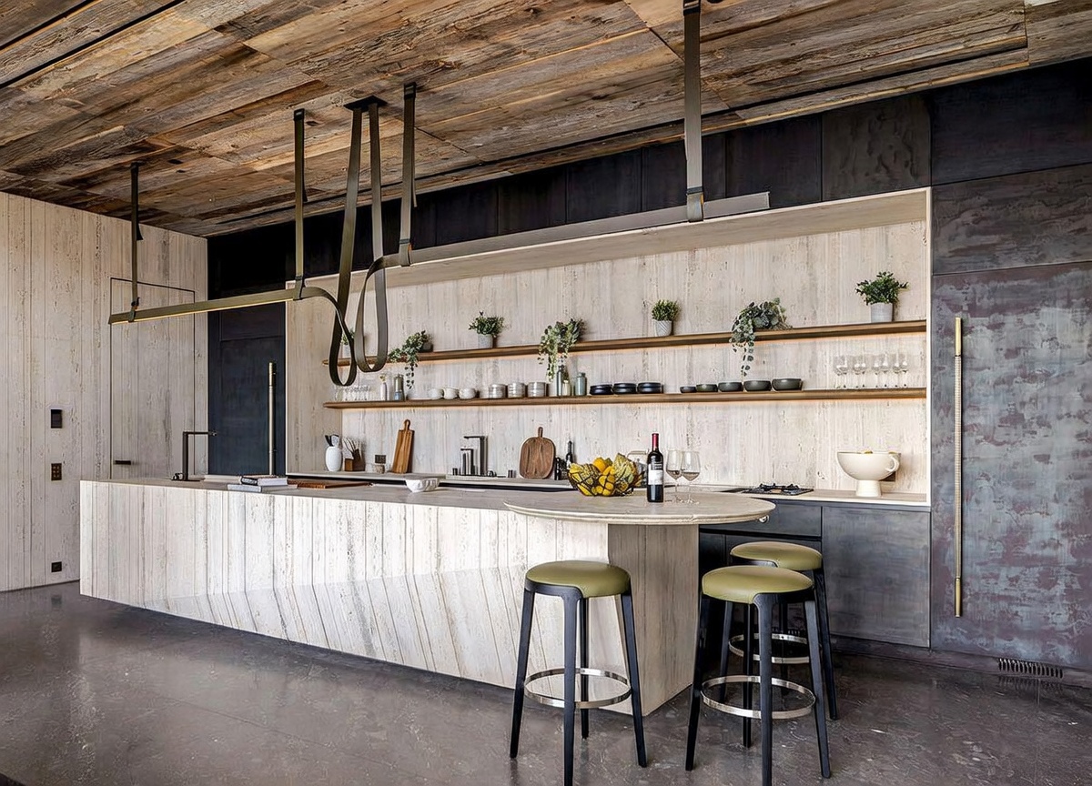 This sophisticated, modern kitchen features a striking combination of raw, natural materials, including a massive travertine stone island and a rustic reclaimed wood ceiling. The space is accented by dark, industrial-style cabinetry, open shelving with curated decor, and unique suspended lighting fixtures. The perspective is a wide-angle shot from the floor level, emphasizing the expansive island and the seamless blend of luxury and organic textures.