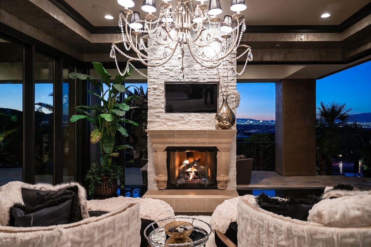 This luxurious living space features a prominent stone-clad chimney breast centered between large glass walls that open to a panoramic city view at dusk. A grand crystal chandelier hangs above a cozy seating area with plush, textured armchairs and a central glass-topped coffee table. The room seamlessly blends indoor comfort with an expansive outdoor terrace, creating an elegant and sophisticated atmosphere.