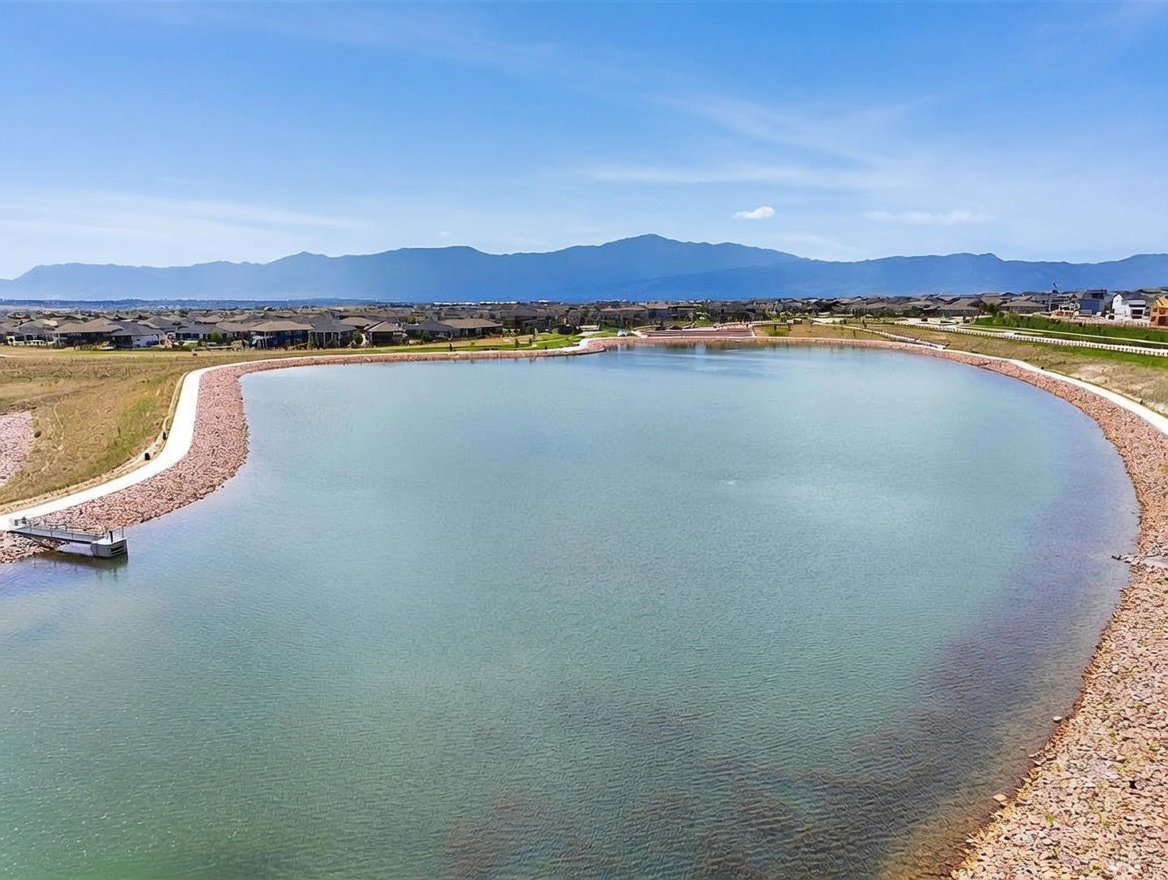 This aerial view captures a large, calm retention pond surrounded by a walking path and rocky shoreline. In the background, a residential neighborhood stretches toward a scenic mountain range under a bright, partly cloudy sky. The perspective provides a clear sense of the community's open space and natural surroundings.