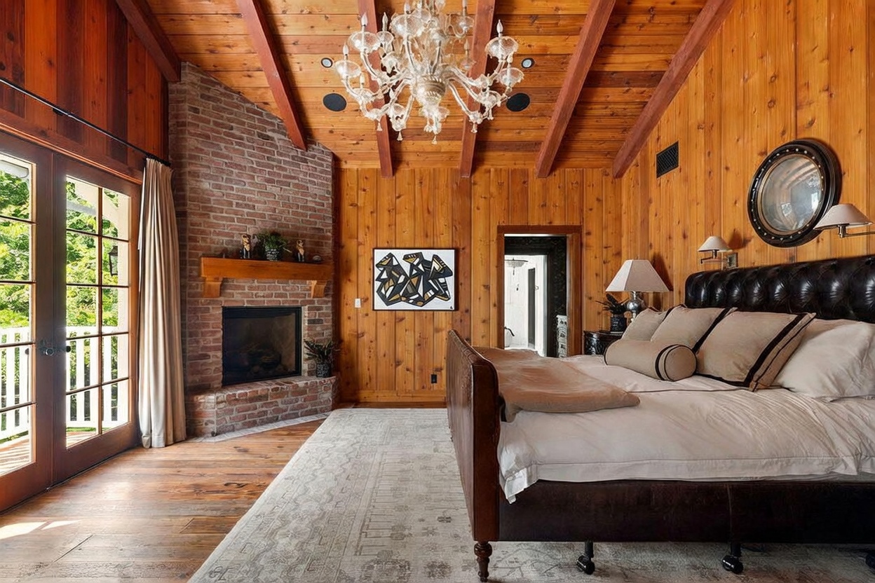 This primary bedroom features warm wood-paneled walls and a vaulted ceiling with exposed beams, creating a rustic and cozy atmosphere. A large brick fireplace with a wooden mantel serves as a focal point, while a leather-tufted bed and a crystal chandelier add a touch of luxury. The room opens to a balcony through glass doors, and a patterned area rug anchors the space, which leads into an adjacent bathroom.