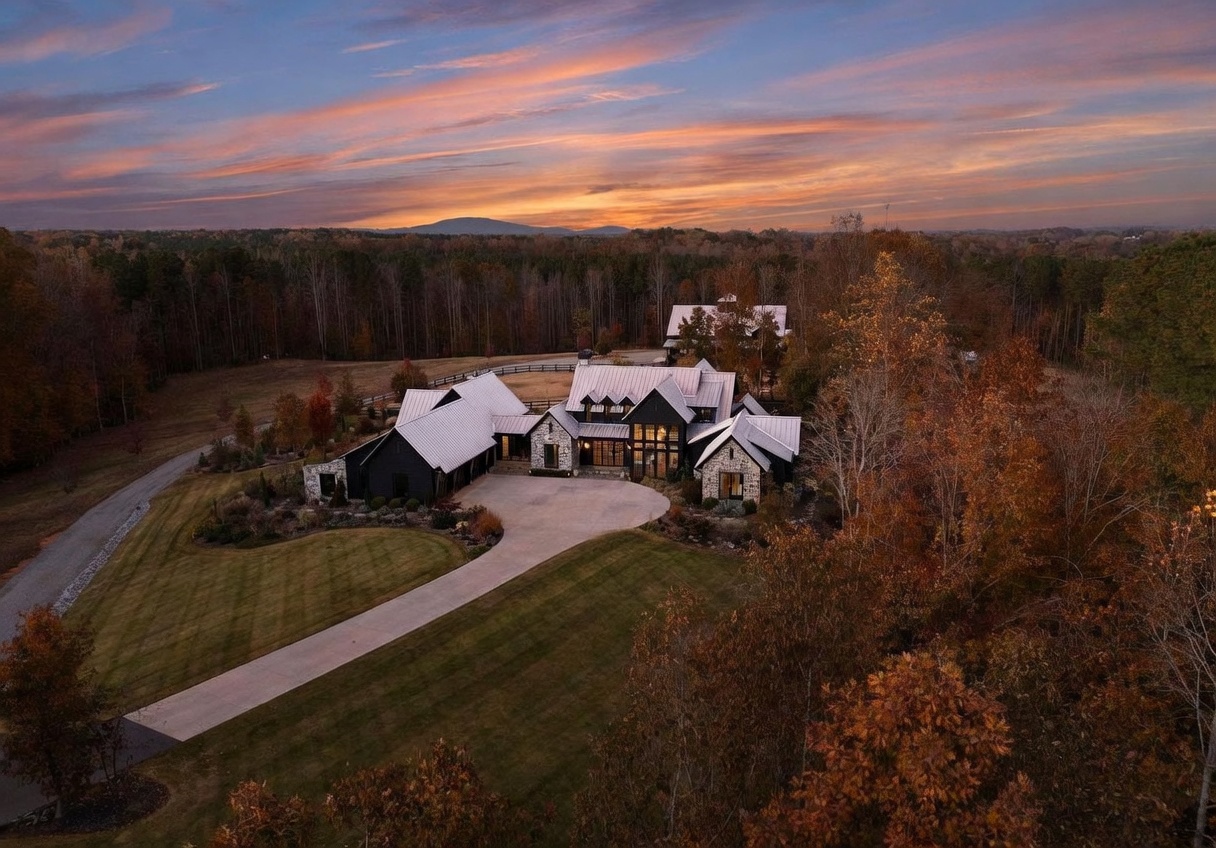 This stunning aerial view captures a modern farmhouse-style estate set against a vibrant sunset sky. The home features a complex roofline with metal roofing, a mix of dark siding and stone accents, and a large circular driveway leading to the main entrance. Surrounded by mature trees and expansive lawns, the property exudes a sense of luxury and seclusion in a picturesque rural setting.
