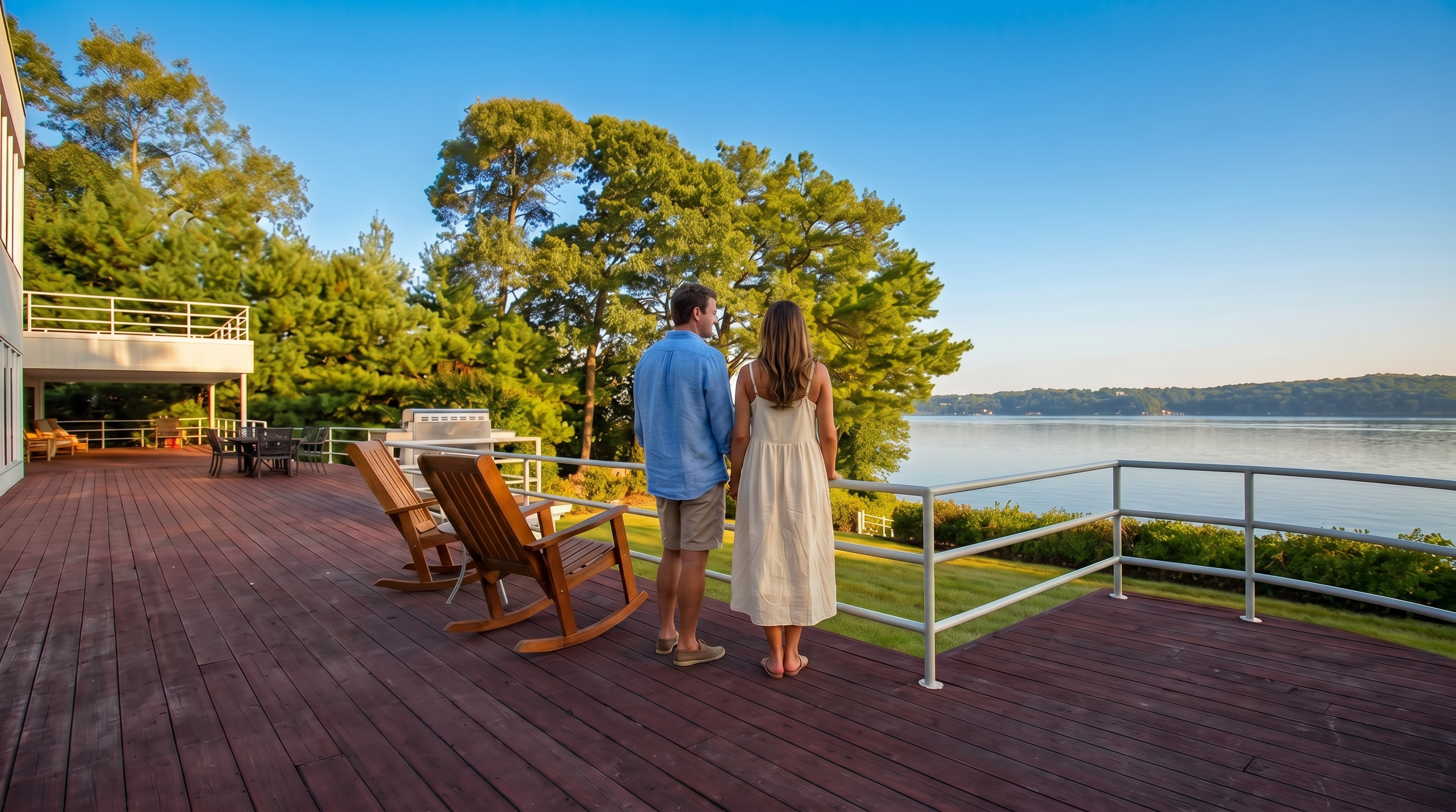 This spacious, elevated wooden deck offers a prime vantage point with stunning, panoramic views of the water. Featuring simple rod railings and a pair of outdoor rocking chairs, the deck provides a serene and scenic setting for relaxation against a backdrop of lush green trees and a clear blue sky.