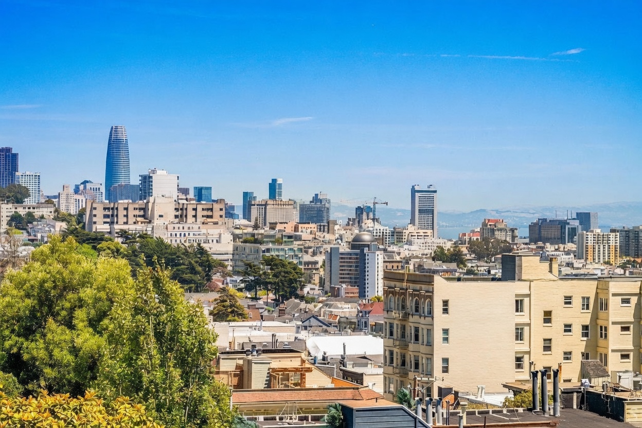 This elevated, wide-angle view captures a sprawling cityscape, highlighting the iconic Salesforce Tower and surrounding urban architecture. The foreground features lush green trees and the rooftops of residential buildings, providing a sense of scale and neighborhood context. The clear blue sky and distant hills create a bright, expansive atmosphere typical of a high-vantage point in San Francisco.