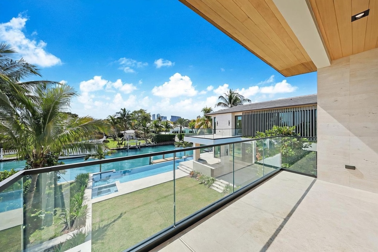This image captures a view from a spacious, covered balcony overlooking a private backyard and a canal. The balcony features a sleek glass railing, light-toned stone flooring, and a warm wood-paneled ceiling, creating a sophisticated outdoor living space. Below, a manicured lawn leads to a swimming pool and a waterfront dock, with a clear blue sky and distant city skyline providing a picturesque backdrop.