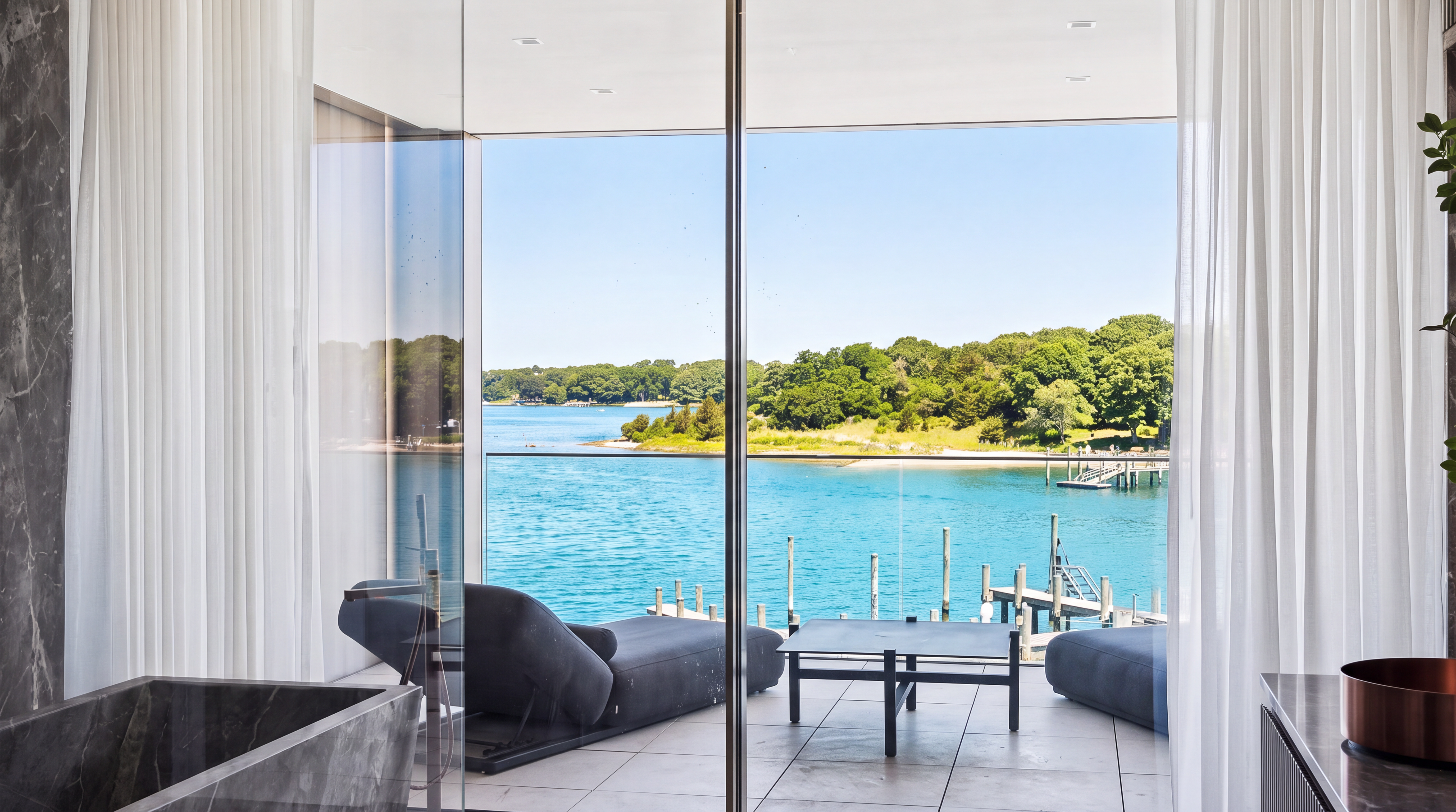 This modern, luxury bathroom features a sleek stone soaking tub and opens seamlessly to a private balcony via floor-to-ceiling glass doors. The balcony offers stunning, unobstructed views of the water, a dock, and the surrounding natural landscape. The space uses high-end materials and clean lines to create a serene, resort-like atmosphere.