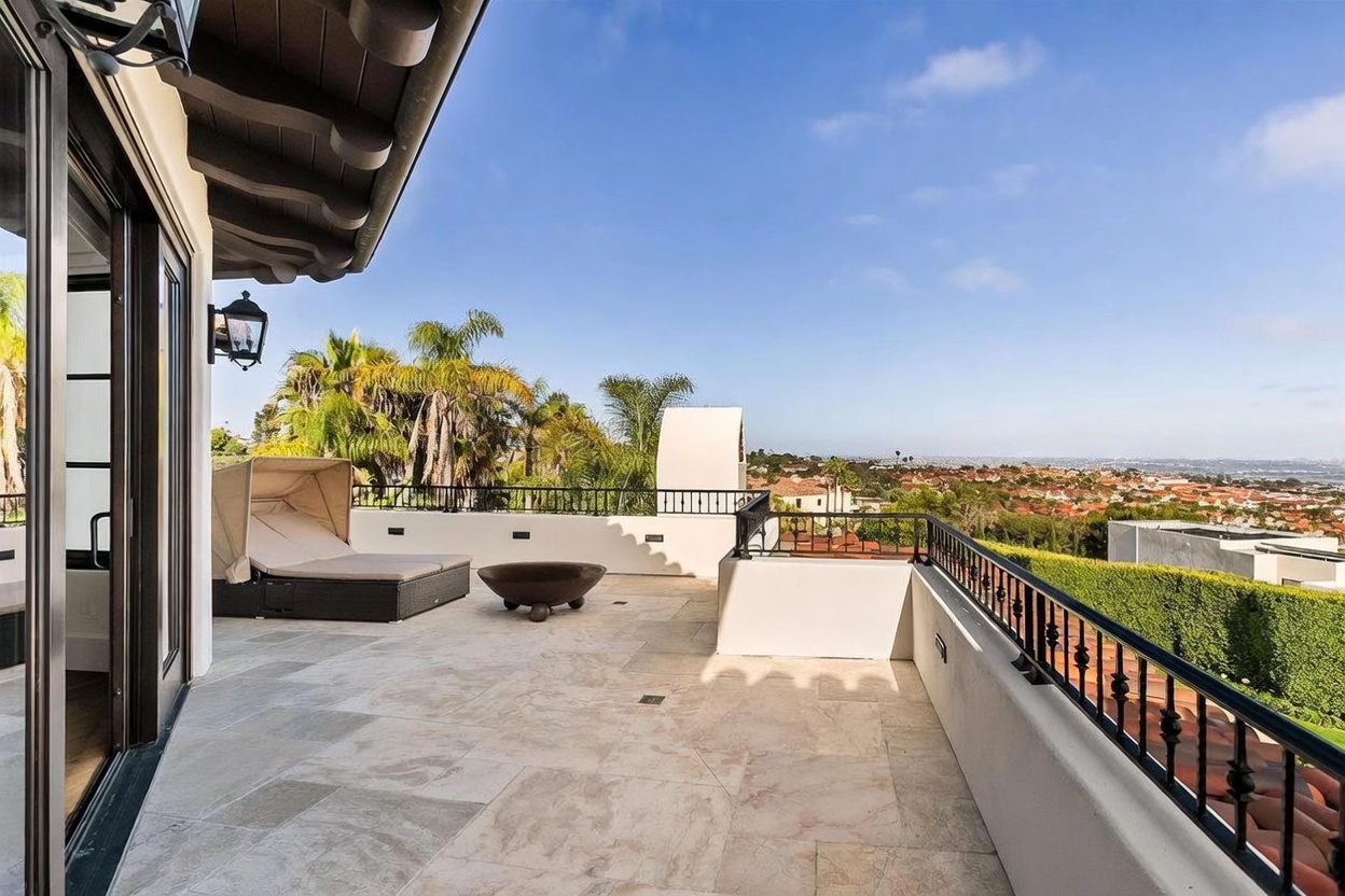 This expansive, elevated balcony features light-toned stone flooring and a black metal railing that overlooks a sprawling suburban landscape. The space is furnished with a modern lounge chair under a canopy and a circular fire pit filled with blue glass, creating an inviting outdoor retreat. The perspective captures the open-air layout, highlighting the seamless transition from the interior through large glass doors to the panoramic views beyond.