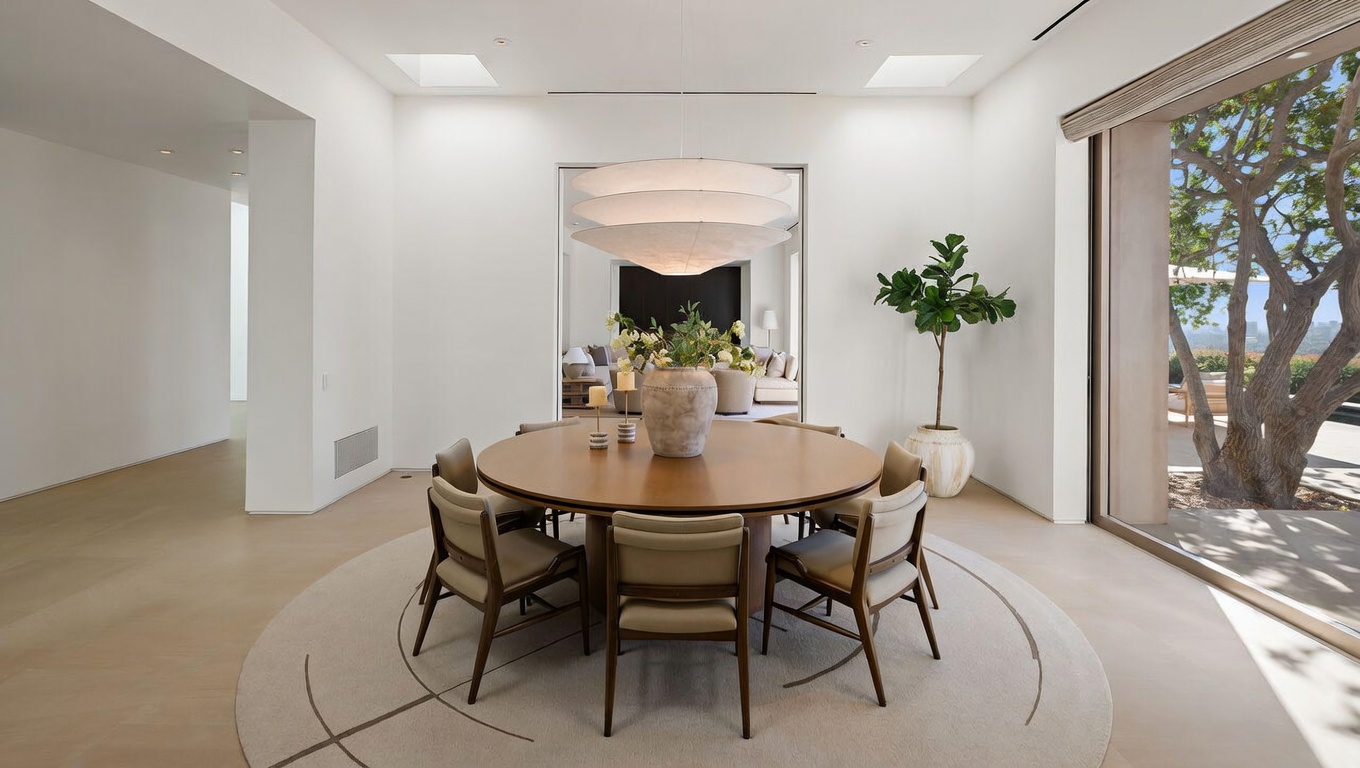 This elegant dining room features a minimalist, modern aesthetic with a large circular wooden table surrounded by six upholstered chairs. A striking, multi-tiered pendant light fixture hangs above the table, while a large window provides a view of a mature tree and outdoor landscape. The room is bright and airy, with clean white walls and a neutral-toned circular rug that anchors the dining area.