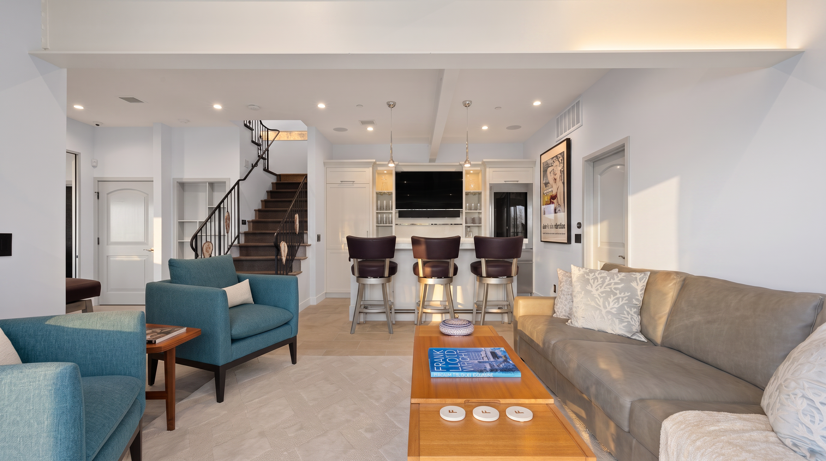 This open-concept living area features a comfortable seating arrangement with a neutral-toned sofa and two vibrant teal armchairs centered around a light wood coffee table. In the background, a built-in bar area with dark stools and a television creates a functional entertainment space, while a staircase with a decorative metal railing leads to an upper level. The room is bright and airy, defined by a prominent white structural beam overhead and a neutral color palette that creates a welcoming, modern atmosphere.