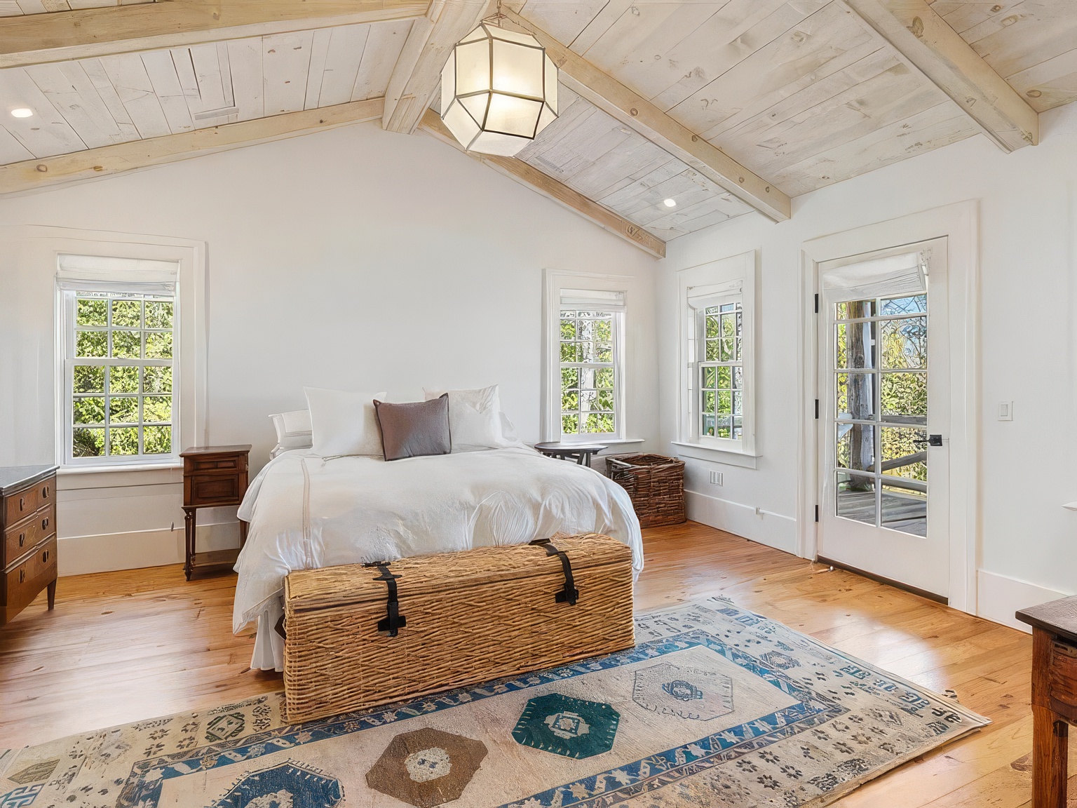 This bright and airy primary bedroom features a vaulted ceiling with exposed light wood beams and a central geometric pendant light. The room is furnished with a bed dressed in white linens, a woven storage chest at the foot, and a patterned area rug over light wood flooring. Multiple windows and a glass-paned door provide ample natural light and views of the surrounding greenery.