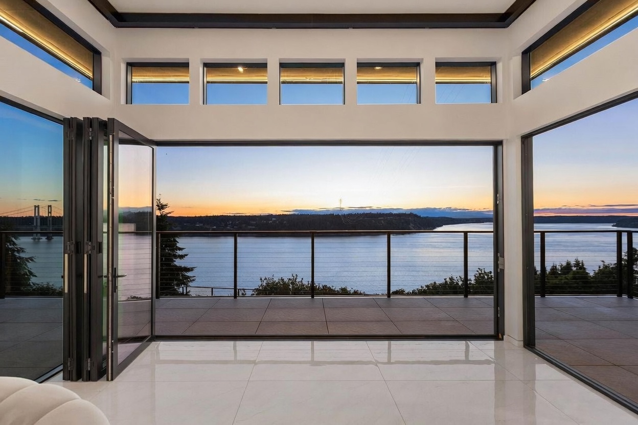 This interior space features expansive floor-to-ceiling glass walls that fold open to reveal a seamless transition to a covered balcony overlooking a serene body of water at sunset. The room is characterized by a minimalist aesthetic with glossy, light-colored tile flooring and a series of clerestory windows that enhance the natural light. The perspective is centered, drawing the eye directly through the open glass panels toward the distant bridge and horizon.