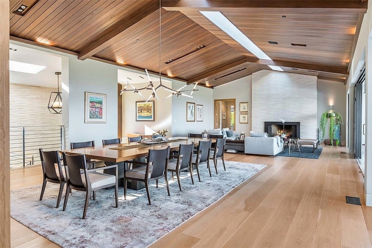 This expansive open-concept living and dining area features a vaulted wood-paneled ceiling and light-toned wood flooring. A long dining table serves as the centerpiece, complemented by a modern chandelier, while a stone-clad chimney breast anchors the adjacent living space. The room is bathed in natural light from large windows and a skylight, creating an airy and sophisticated atmosphere.