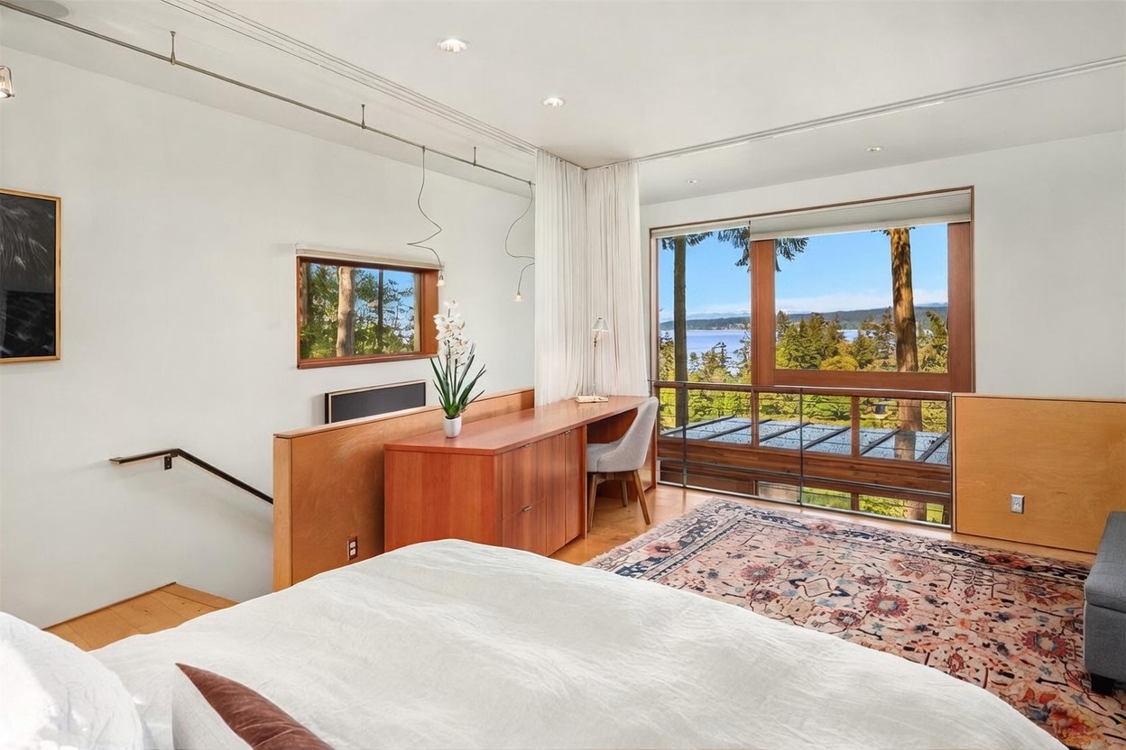 This primary bedroom features a contemporary design with a built-in wooden desk area and a staircase leading to a lower level. Large windows provide an expansive view of the surrounding trees and water, while a patterned area rug adds texture to the light wood flooring. The space is bright and airy, emphasizing a seamless connection between the interior living area and the natural landscape outside.