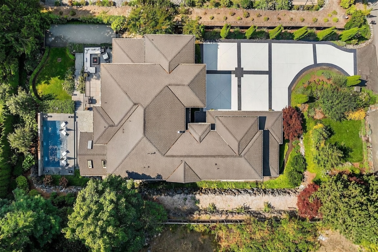 This high-angle aerial view captures a sprawling luxury residence with a complex, multi-gabled roofline and a large, geometric-patterned concrete driveway. The property features a private swimming pool on the left side, adjacent to a stone-paved patio area with outdoor seating. Lush landscaping, mature trees, and manicured lawns surround the home, creating a private and serene estate atmosphere.