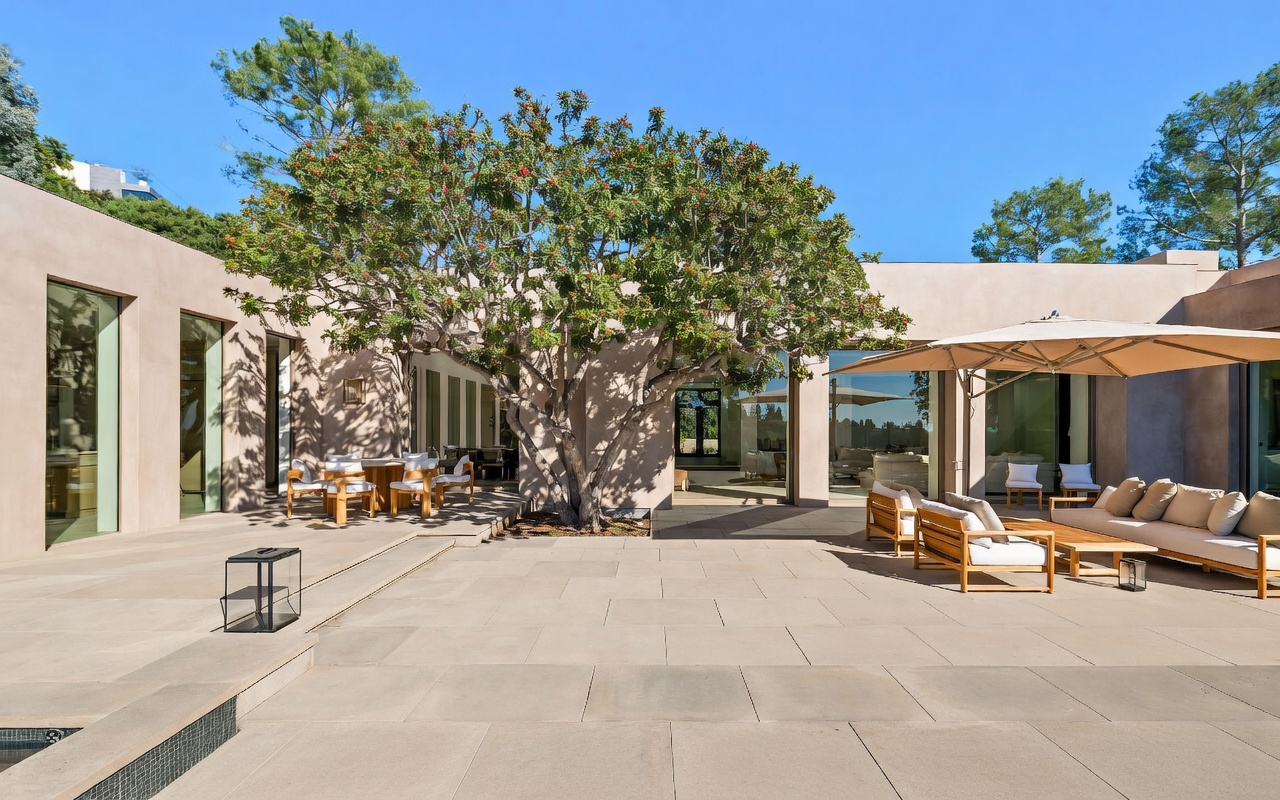 This expansive, modern patio area features a seamless indoor-outdoor flow with large glass sliding doors and light-toned stone pavers. A mature, leafy tree serves as a central focal point, surrounded by comfortable outdoor lounge furniture and a dining set under a large umbrella. The scene is captured from a low, eye-level perspective, emphasizing the serene, resort-like atmosphere and the adjacent swimming pool.