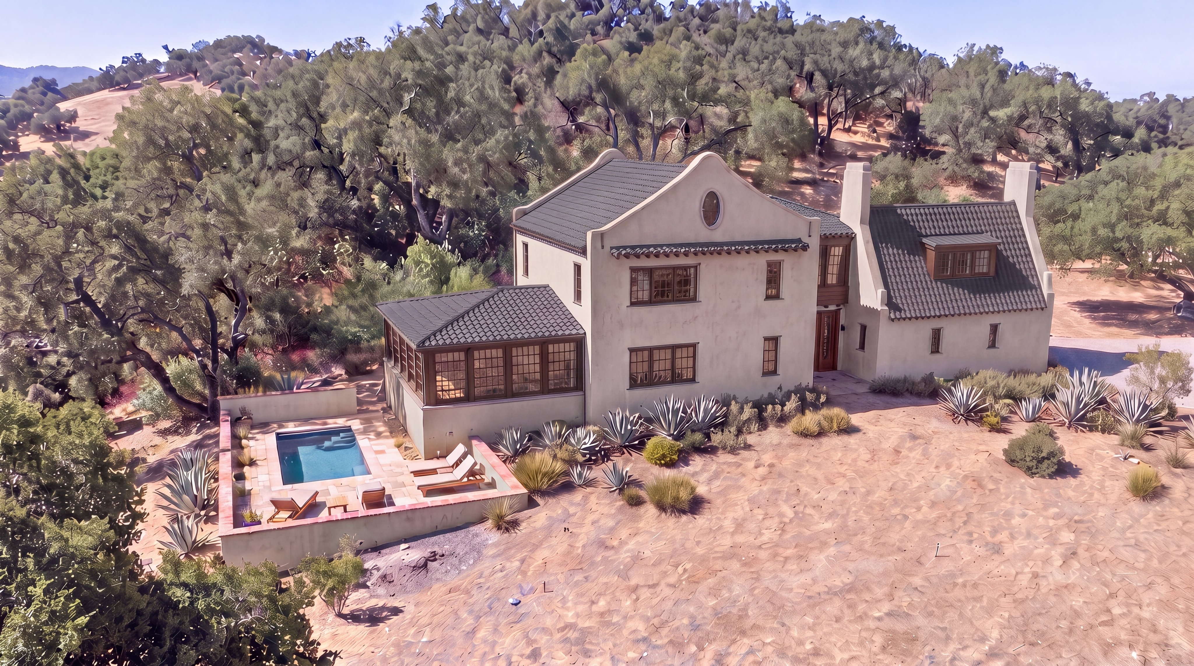 This stunning aerial view captures a Mediterranean-style home nestled into a hillside landscape, highlighting its integration with the natural, arid environment. The property features a tiered stone patio with a swimming pool and lounge area, contrasting beautifully with the surrounding trees and shrubs. Its architectural charm is defined by a multi-sectioned roofline, soft stucco exterior, and abundant windows that suggest a sophisticated and tranquil retreat.