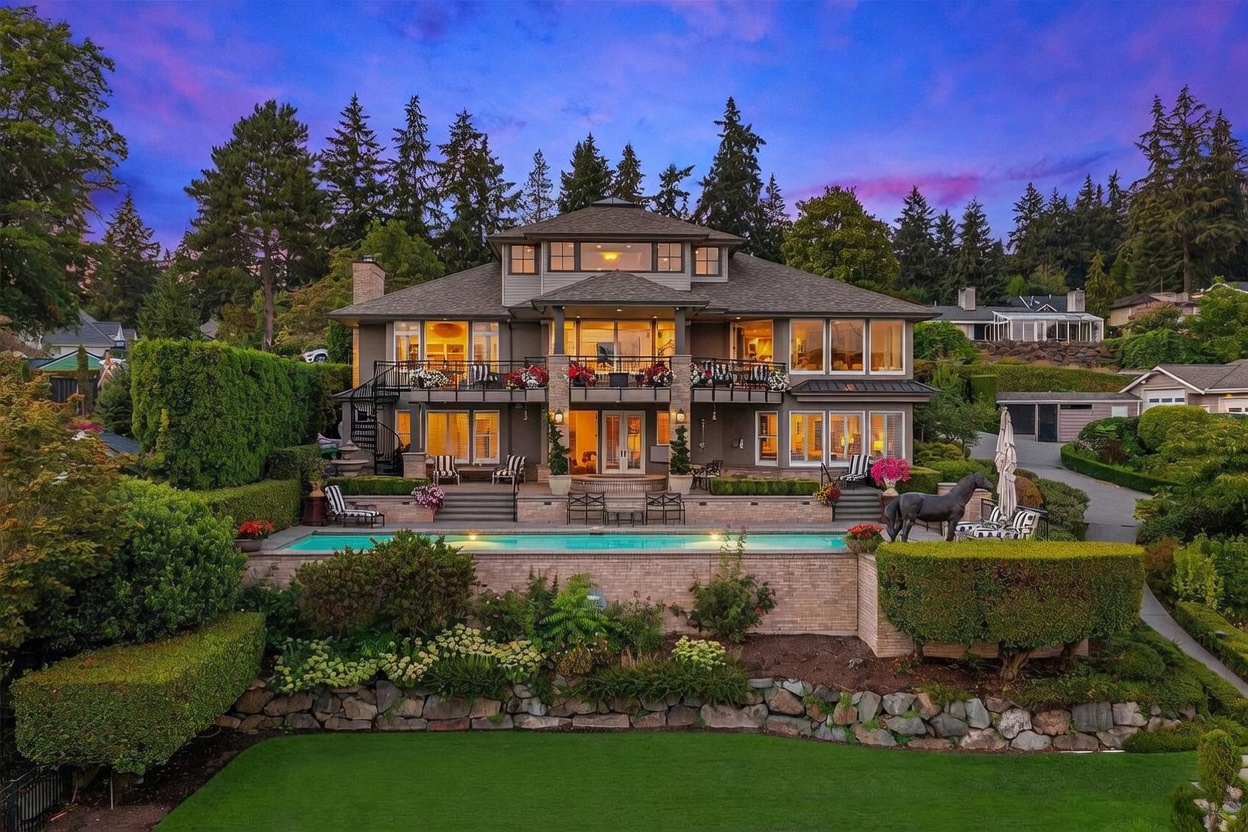 This stunning rear view of a multi-story luxury residence showcases a sprawling terrace with a swimming pool, manicured hedges, and lush landscaping. The home features a symmetrical design with large windows, a spacious balcony, and a spiral staircase leading to the upper level. The twilight sky provides a dramatic backdrop, highlighting the warm interior lighting and the elegant, tiered outdoor living spaces.