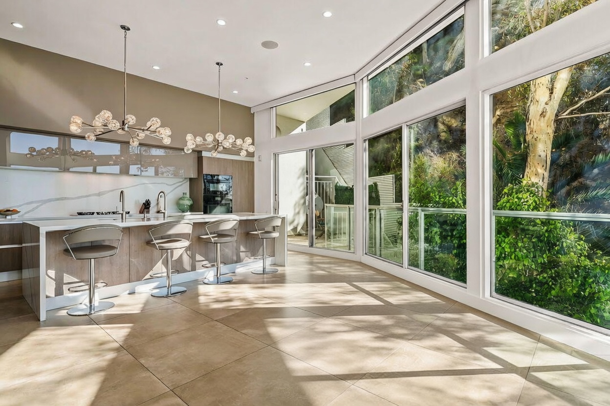 This modern, open-concept kitchen and dining area features a large island with bar seating and a glass-topped dining table. The space is defined by its expansive floor-to-ceiling windows that provide a seamless connection to the lush, wooded exterior. Elegant, multi-bulb pendant lights hang above the island, complementing the sleek cabinetry and neutral tile flooring.
