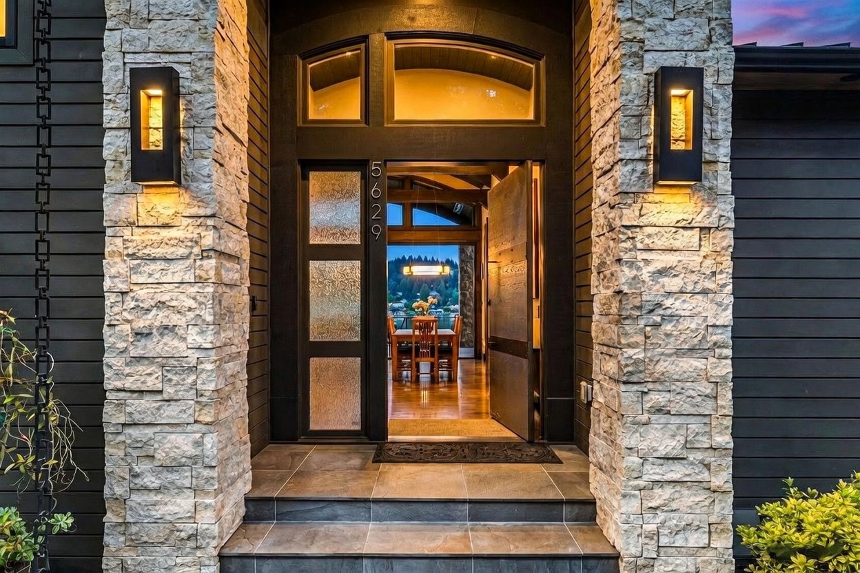 This inviting front entryway features a grand door with frosted glass panels and a transom window, framed by textured stone pillars and dark horizontal siding. Two modern, rectangular wall sconces provide warm illumination, highlighting the stone texture and the transition into the home. Through the open door, a glimpse of the interior dining area and a scenic outdoor view creates a welcoming and sophisticated first impression.