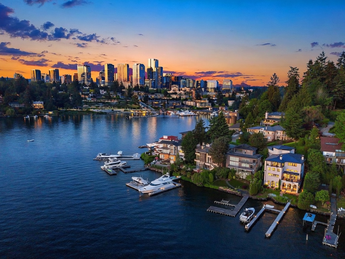 This stunning aerial perspective captures a luxury waterfront property at sunset, showcasing its private dock with multiple boats and direct access to the lake. The multi-level residence features expansive windows and tiered outdoor spaces that overlook the calm water, with the vibrant city skyline glowing in the distance. The composition highlights the prime location, blending serene natural surroundings with the convenience of urban proximity.