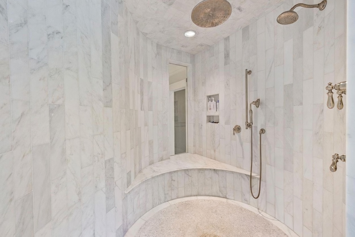 This spacious walk-in shower features light-toned stone wall tiles arranged in a vertical pattern and a curved built-in bench. The shower is equipped with a large overhead rainfall showerhead, a separate handheld sprayer on a sliding bar, and multiple wall-mounted controls. A recessed niche provides convenient storage for toiletries, and the floor is finished with small, circular mosaic tiles.