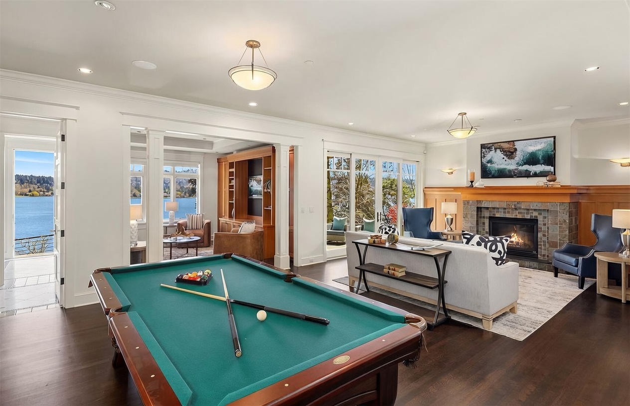 This spacious, open-concept living area features a billiard table in the foreground and a cozy seating area with a fireplace in the background. The room is characterized by dark hardwood floors, elegant crown molding, and large glass doors that provide a stunning view of the lake. The overall aesthetic is sophisticated and inviting, blending recreational space with a comfortable living environment.