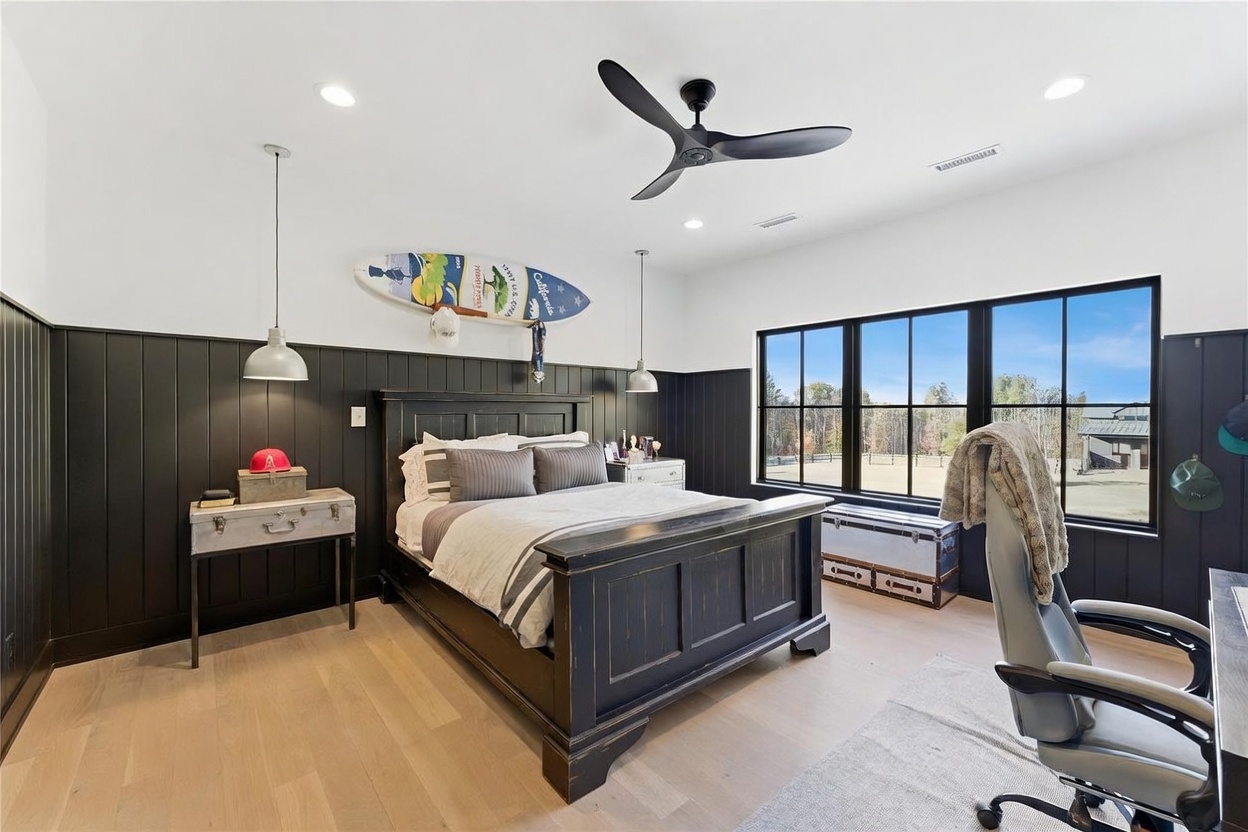 This bedroom features a bold aesthetic with dark, vertical wood paneling on the lower walls contrasted against crisp white upper walls and light wood flooring. A dark, traditional-style bed frame serves as the centerpiece, accented by a decorative surfboard mounted above the headboard and industrial-style pendant lighting. Large windows with black frames provide ample natural light and a view of the outdoor landscape, creating a balanced, modern-masculine atmosphere.