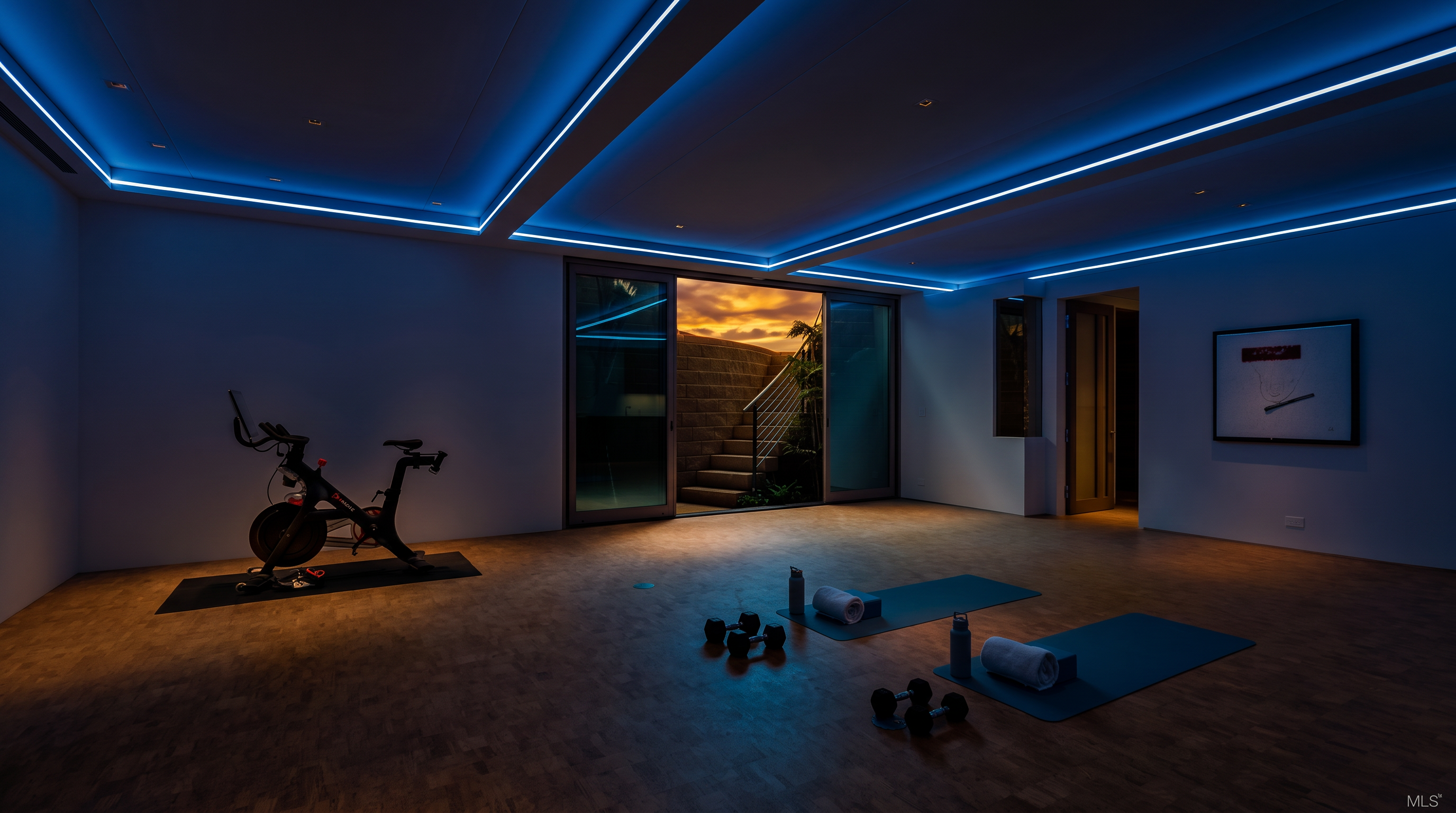 This spacious home gym features light-toned cork flooring and a minimalist aesthetic, highlighted by recessed ceiling lighting with blue accent strips. A stationary exercise bike sits on a mat to the left, while two yoga mats with dumbbells and towels are arranged on the floor in the center. Large sliding glass doors open to an outdoor area with a curved stone staircase, providing a seamless transition between indoor and outdoor fitness spaces.