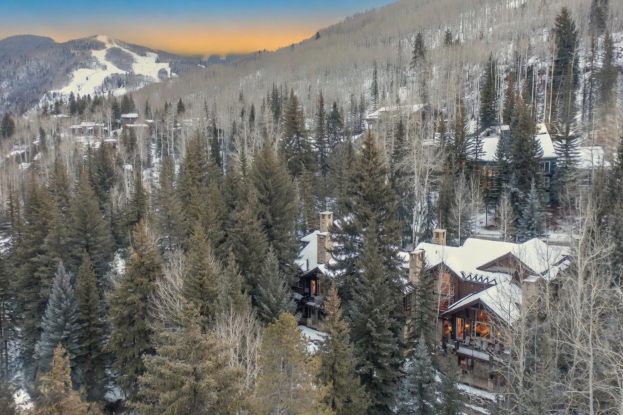 This aerial view captures a large, multi-story mountain home nestled within a dense forest of evergreen and aspen trees. The architecture features a rustic aesthetic with dark wood siding, stone accents, and multiple gabled rooflines covered in a light dusting of snow. In the background, a snow-covered ski slope is visible against a soft, sunset-lit sky, emphasizing the property's prime location in a winter resort setting.