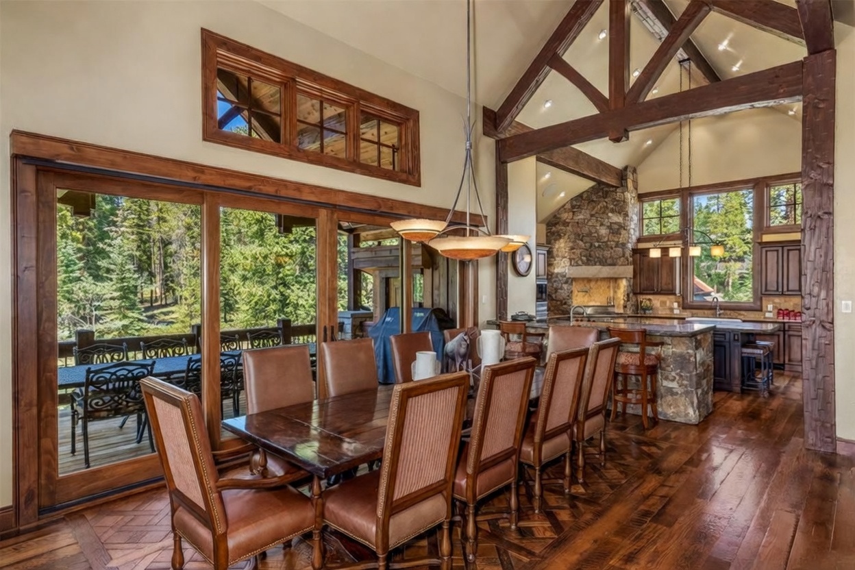 This elegant dining room features a large wooden table surrounded by upholstered chairs, set against a backdrop of expansive glass sliding doors that open to a deck. The space is defined by rich wood flooring in a herringbone pattern and exposed timber beams that draw the eye upward toward the vaulted ceiling. In the background, a stone-clad chimney breast anchors the adjacent kitchen area, which is equipped with a stone-topped island and bar seating.