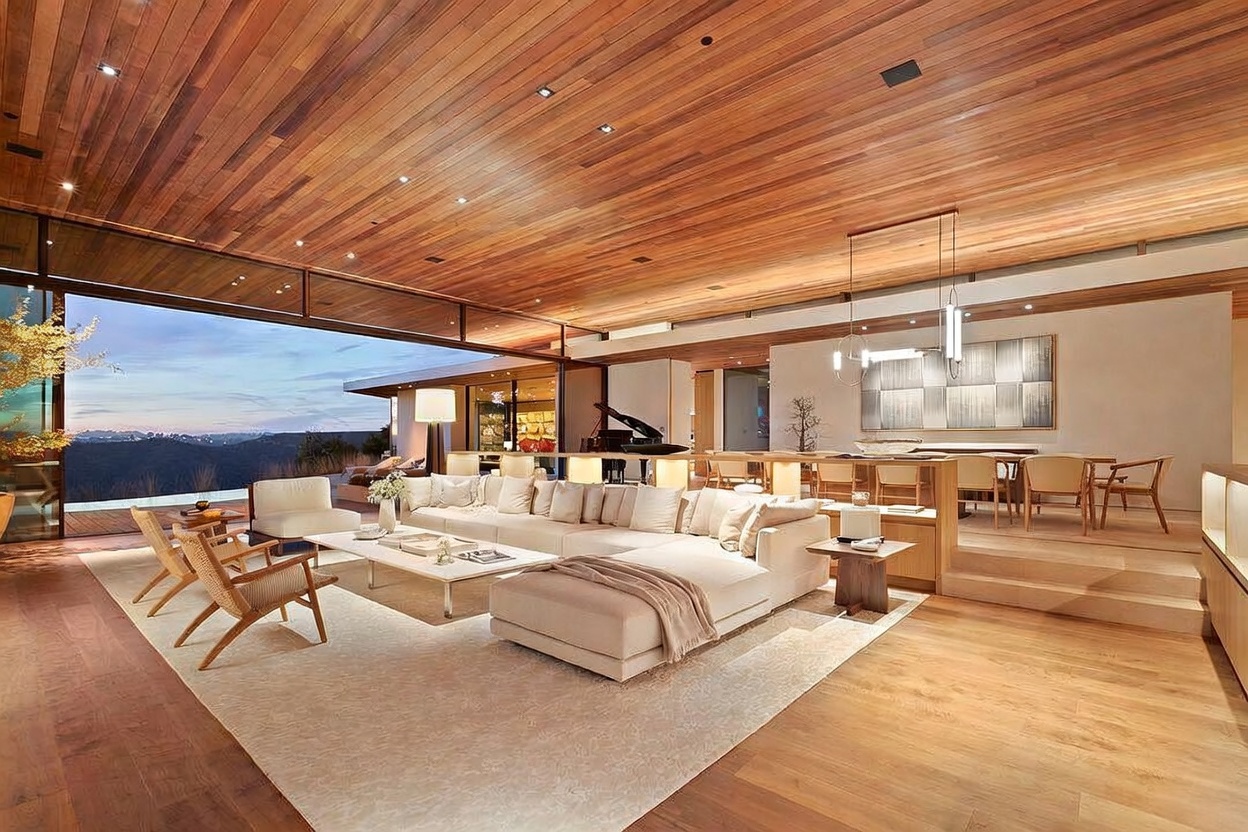 This expansive, open-concept living area features a stunning wood-paneled ceiling and floor-to-ceiling glass walls that seamlessly blend the interior with the outdoor landscape. The space is anchored by a large, neutral-toned sectional sofa and a minimalist coffee table, creating a sophisticated and airy atmosphere. A raised dining area sits in the background, while the warm wood tones and recessed lighting provide a luxurious, modern aesthetic.