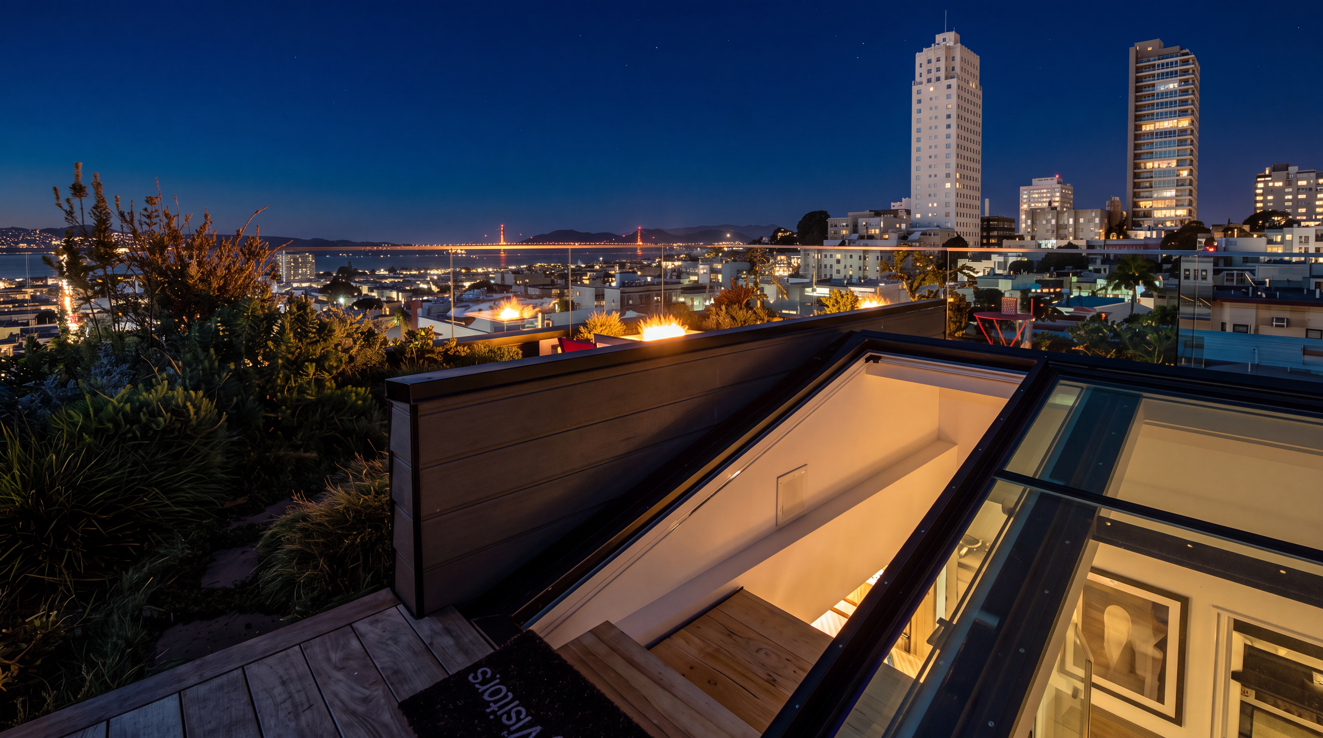 This stunning rooftop deck offers a contemporary outdoor living experience with panoramic views of the city skyline and the Golden Gate Bridge. The space features modern decking, built-in fire elements, and a retractable glass skylight that provides a seamless transition to the interior. It represents a sophisticated urban retreat, perfect for evening gatherings and enjoying high-end city living.