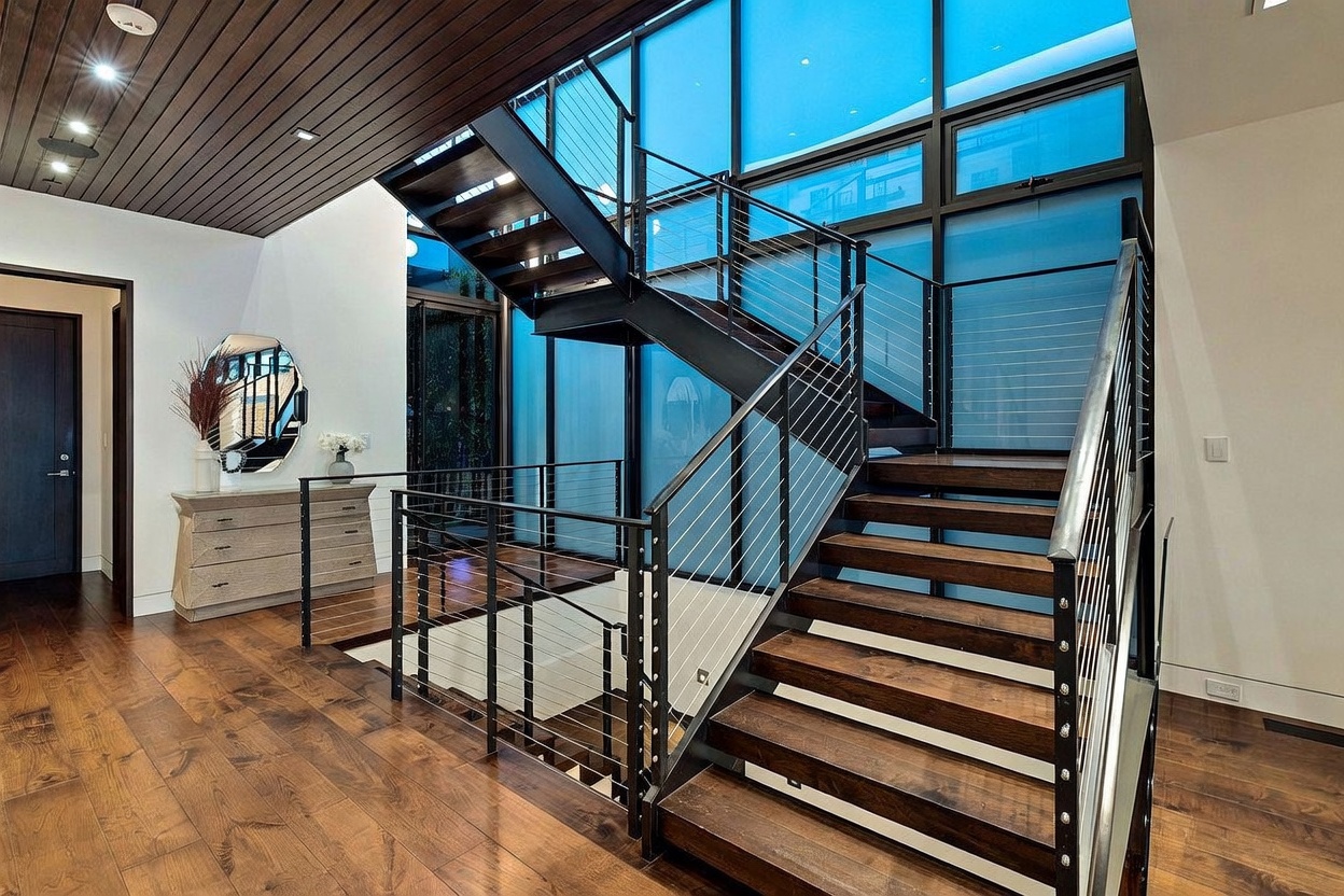 This striking entryway features a dramatic open-riser staircase with dark metal railings and wood treads, serving as the focal point of the space. The area is defined by a high, wood-paneled ceiling and expansive floor-to-ceiling windows that flood the interior with natural light. A sleek console table with a circular mirror sits to the side, enhancing the modern, architectural aesthetic of this grand foyer.
