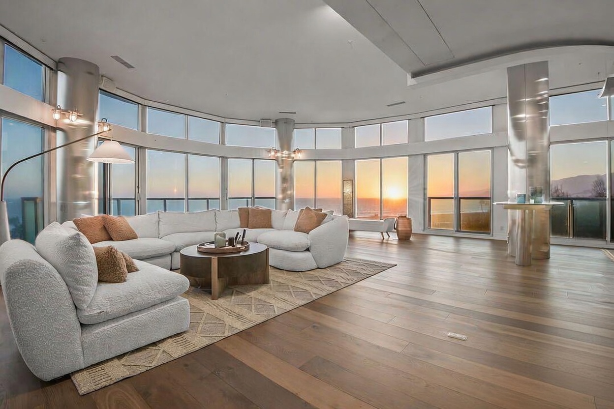 This expansive living room features a large, curved sectional sofa arranged on a textured area rug, centered around a circular coffee table. The space is defined by a sweeping wall of floor-to-ceiling windows that offer panoramic ocean views and a sunset backdrop. Polished metal columns and warm-toned wood flooring enhance the modern, airy aesthetic of this high-end coastal residence.