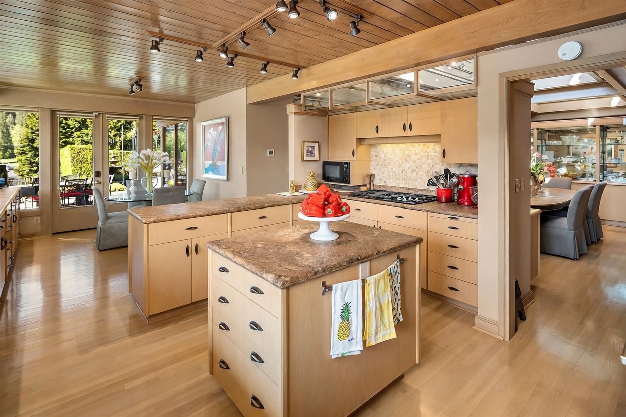 This spacious kitchen features light wood cabinetry, a central island with a stone countertop, and a gas cooktop set into a tiled backsplash. The room is illuminated by track lighting mounted on a wood-paneled ceiling and includes a dining area visible through large windows that look out onto a lush green yard. The perspective is from the center of the kitchen, showcasing an open-concept layout that flows into an adjacent dining space.