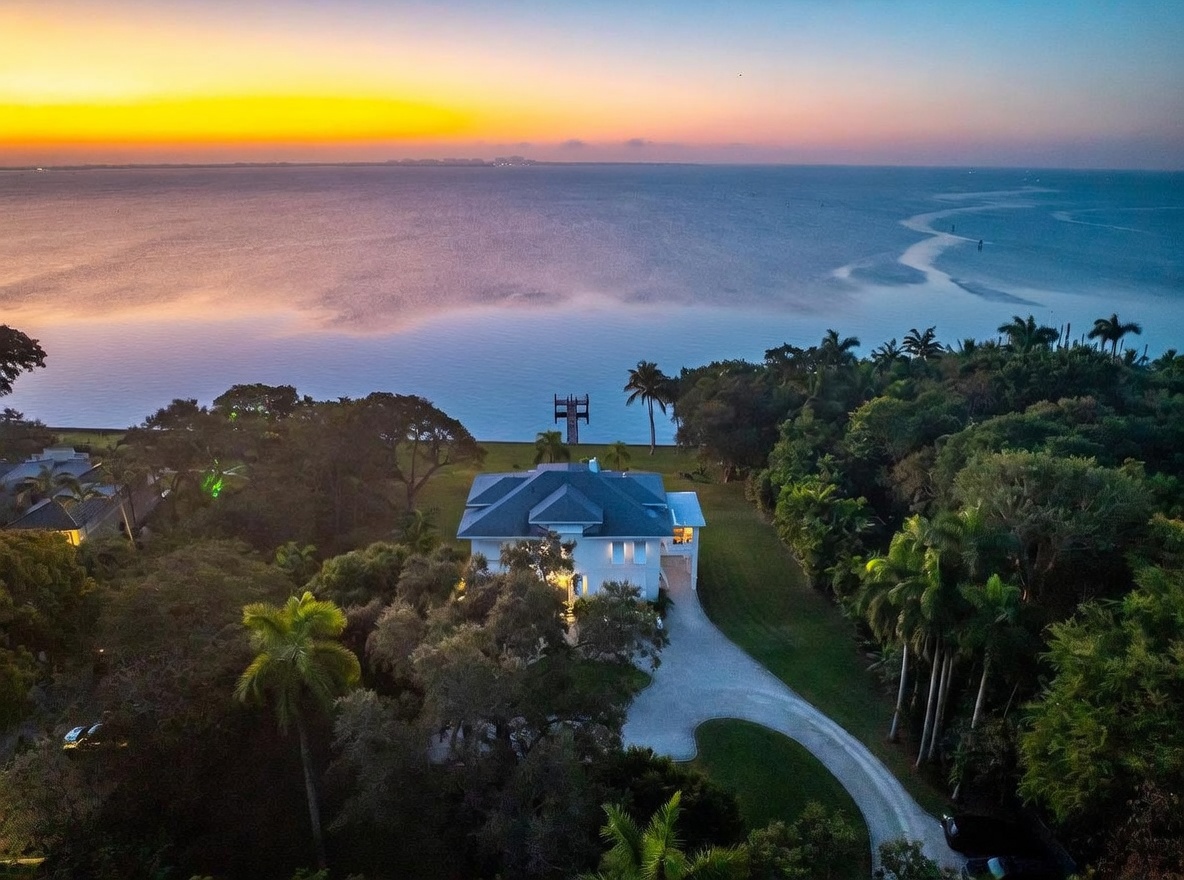This stunning aerial perspective captures a luxurious waterfront estate at sunset, showcasing the property's expansive grounds and direct access to the bay. The white multi-story residence is nestled among lush, mature trees and features a winding driveway that leads to the home's entrance. The vibrant orange and purple hues of the twilight sky reflect beautifully across the calm water, creating a serene and high-end atmosphere.