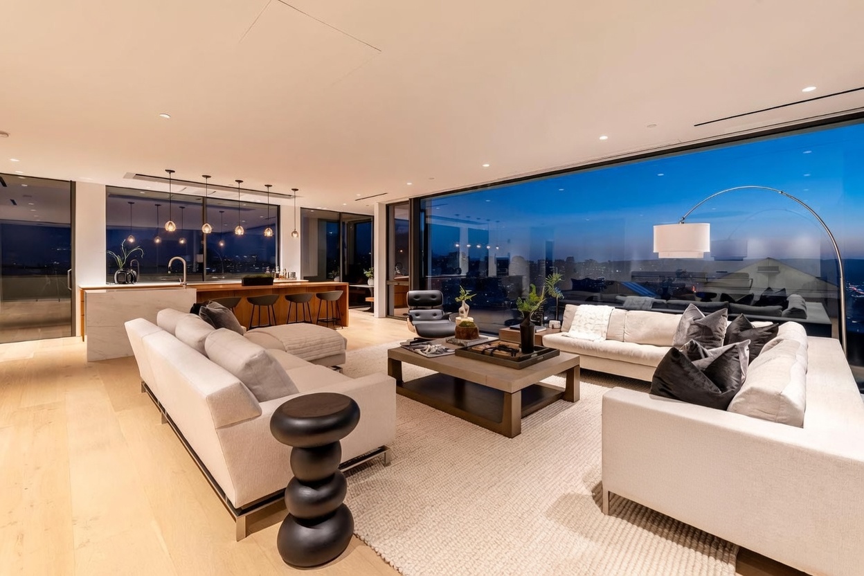 This expansive, open-concept living area features a sophisticated modern aesthetic with light wood flooring and a neutral color palette. The space is anchored by two large sofas facing a central coffee table, complemented by unique sculptural side tables and a tall, arched floor lamp. Floor-to-ceiling glass walls provide a panoramic view of the city skyline at dusk, seamlessly connecting the interior living space to the urban horizon.